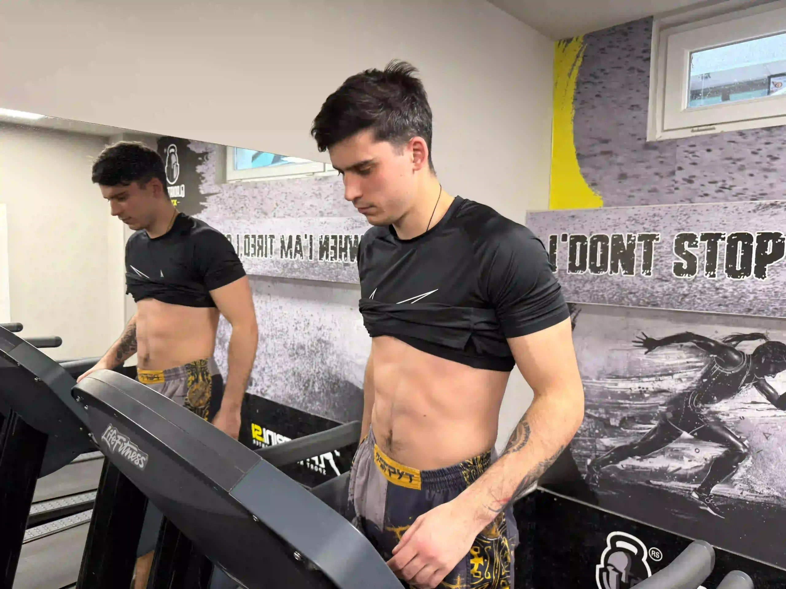 Vanja in a gym standing on a treadmill with his t-shirt pulled up to show his abdominal muscles.