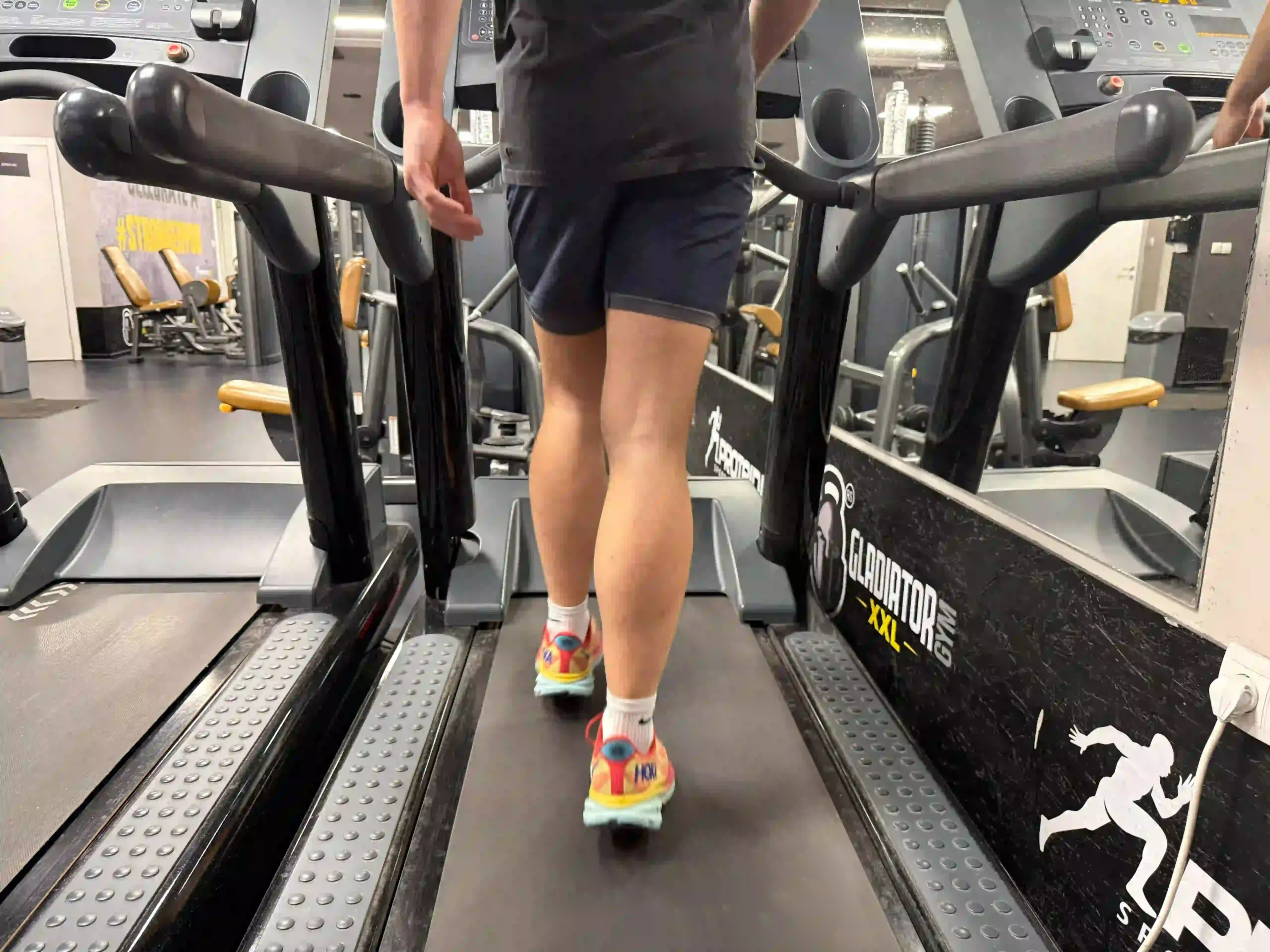 Milutin walking on commercial treadmill in dark tank top, shorts, and colorful running shoes at gym facility with multiple cardio machines