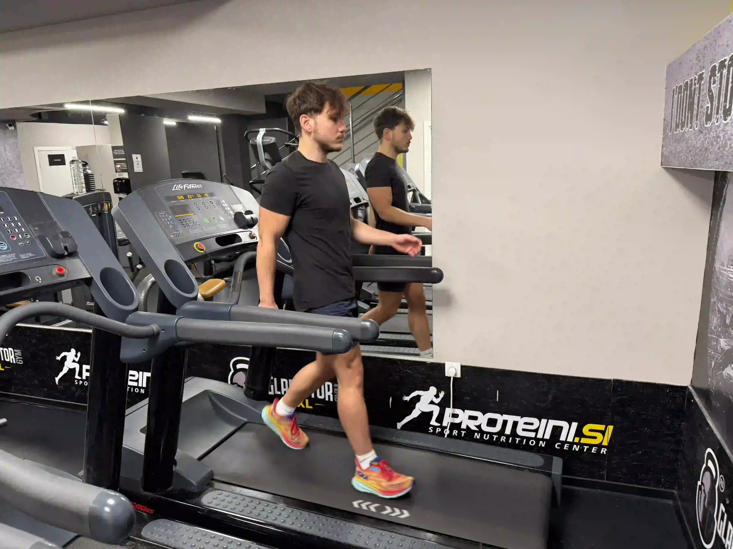 Young man in a black t-shirt walking backward on a treadmill in a gym.