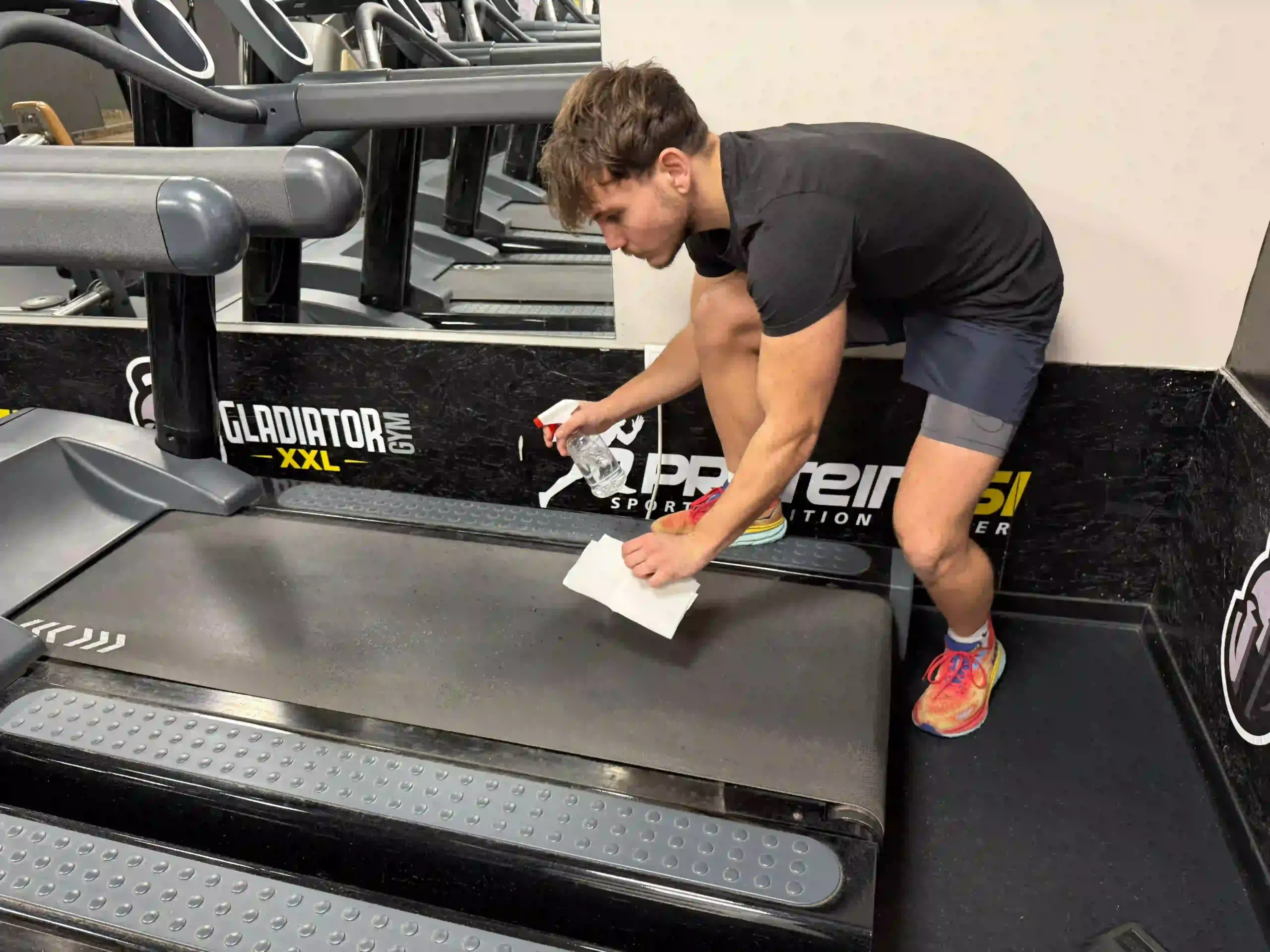 Milutin cleaning a treadmill belt with a spray and cloth to maintain gym hygiene and equipment longevity.