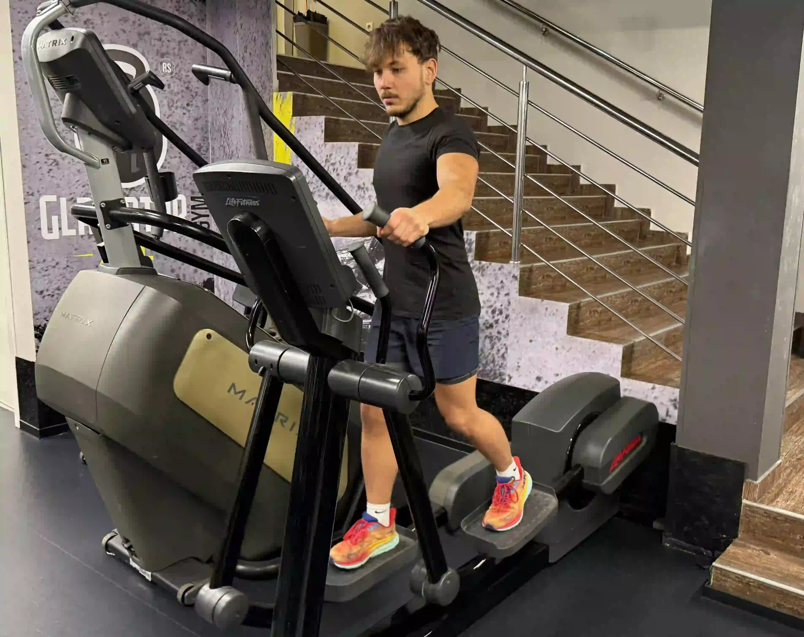 Milutin exercising on elliptical machine in black athletic shirt and shorts with colorful running shoes at gym with staircase in background