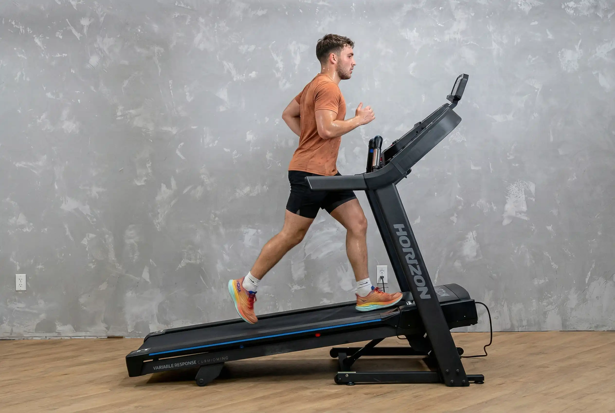 Milutin running on Horizon Fitness 7.0 AT treadmill at incline in orange shirt and black shorts against industrial gray concrete wall