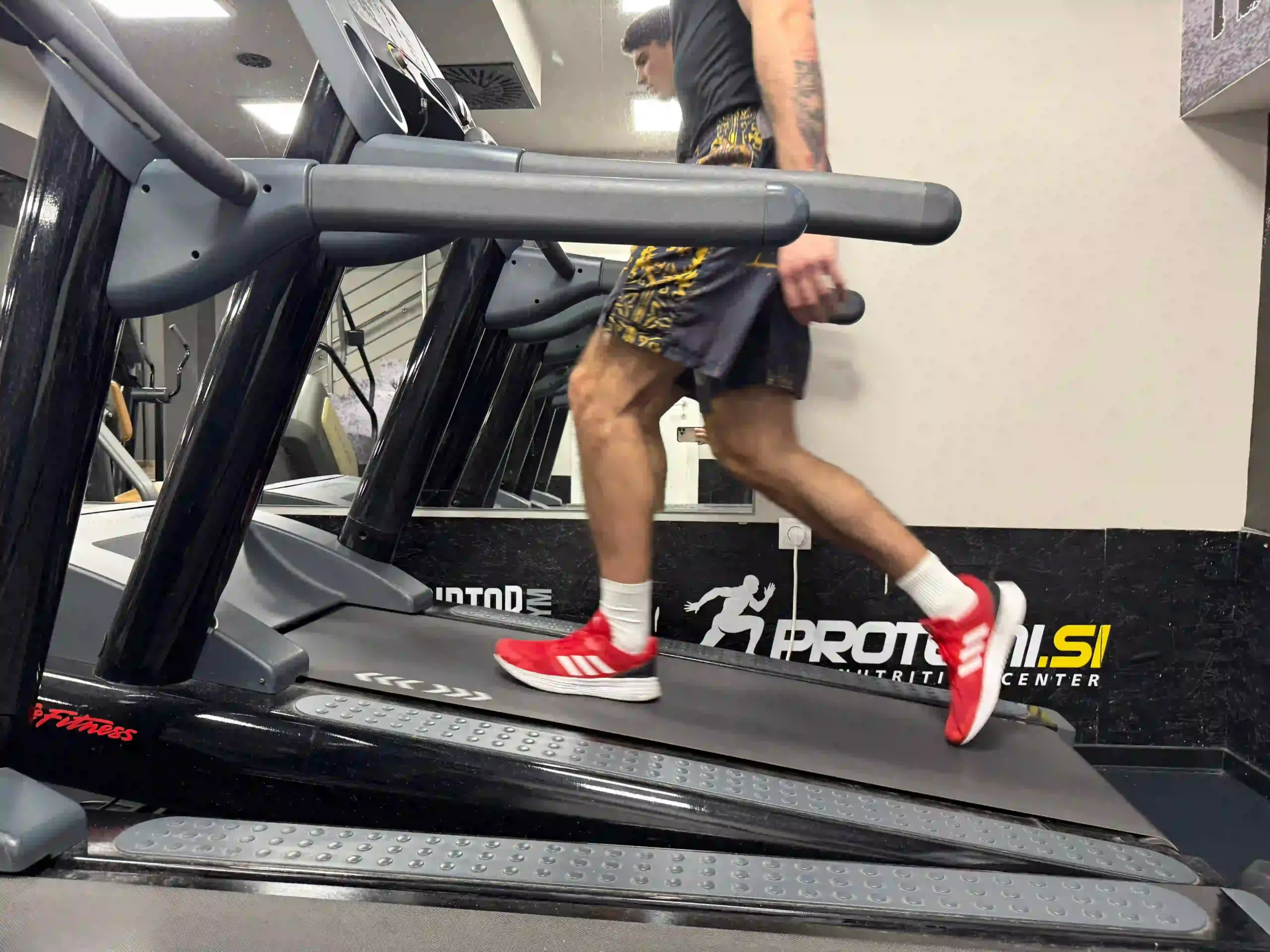 A young man named Vanja wearing a black t-shirt and red sneakers, walking on a treadmill in front of a mirror at the gym.