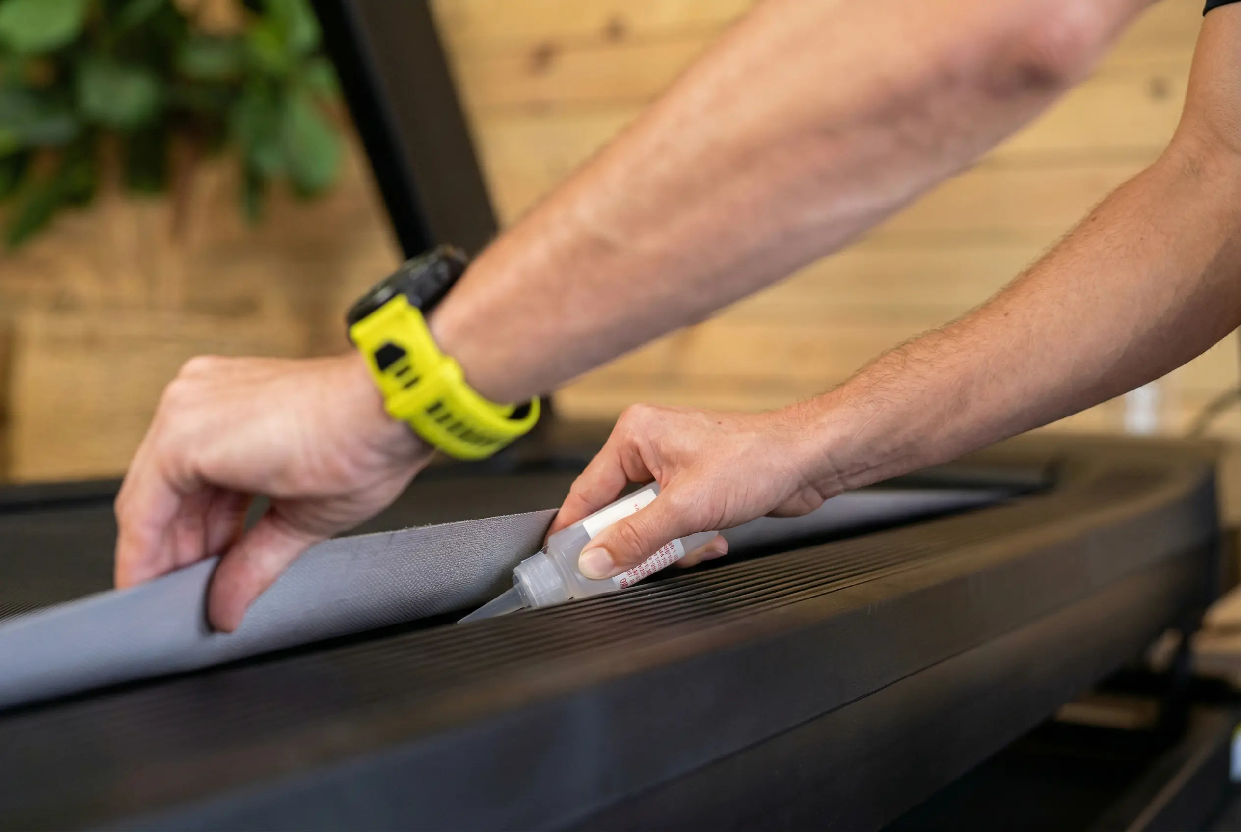Hands applying lubricant underneath treadmill belt on NordicTrack Commercial 1750, lifting edge of running deck with yellow fitness tracker visible on wrist