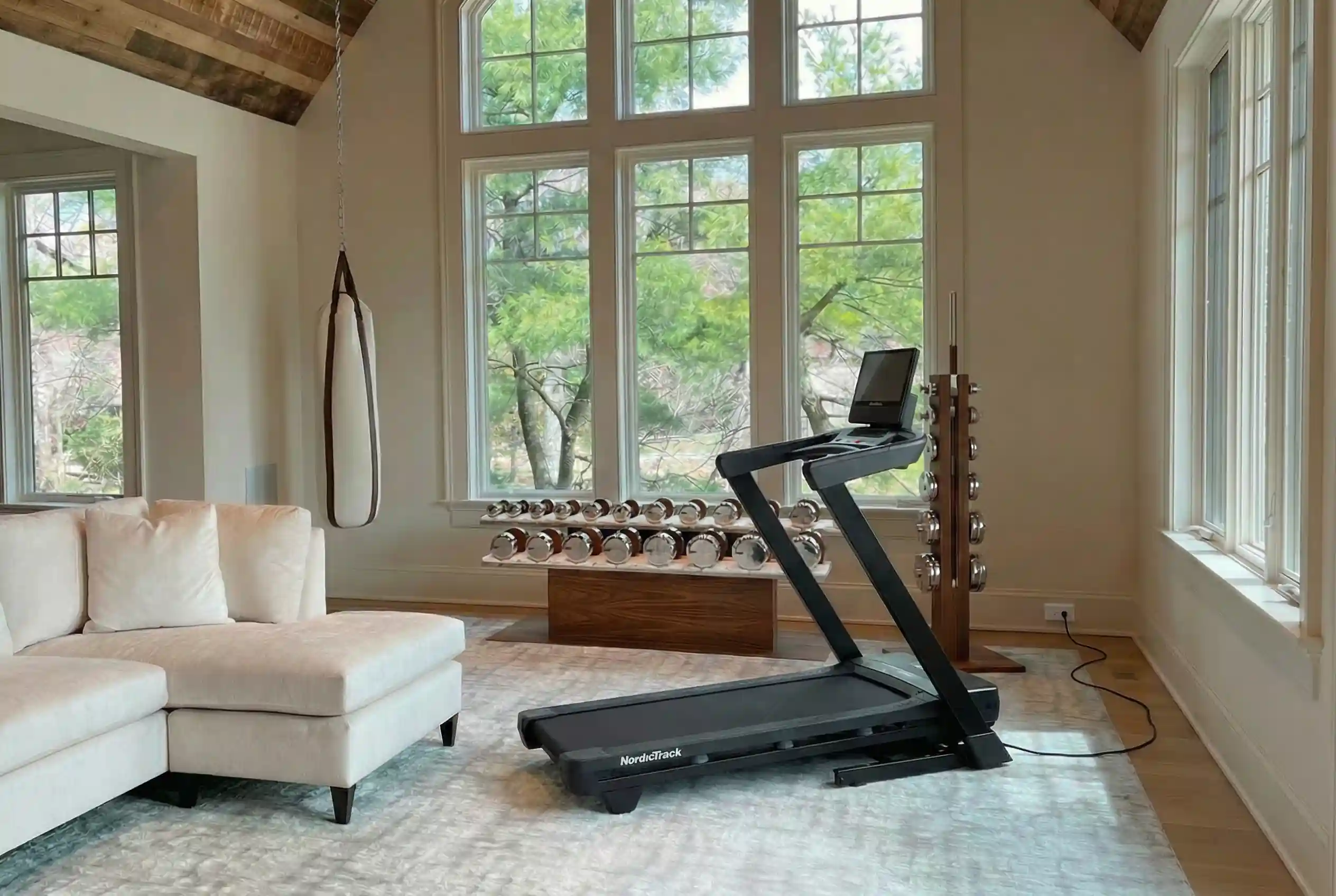 NordicTrack T Series 16 treadmill in luxury home gym with arched windows, exposed beam ceiling, chrome dumbbell set, and white sectional sofa