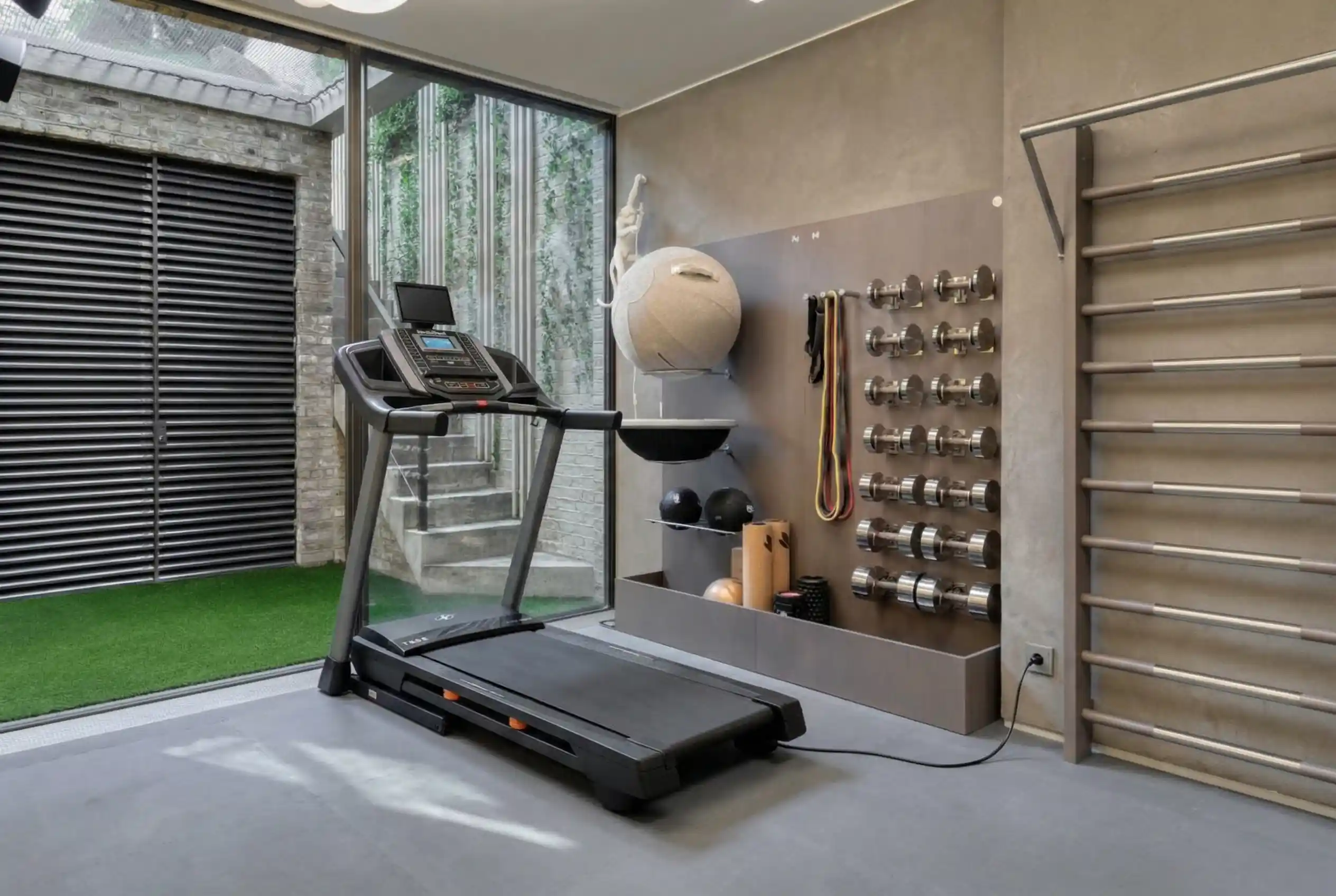 NordicTrack T Series 6.55 treadmill in modern home gym with artificial turf flooring, wall-mounted dumbbell rack, and glass door to outdoor courtyard