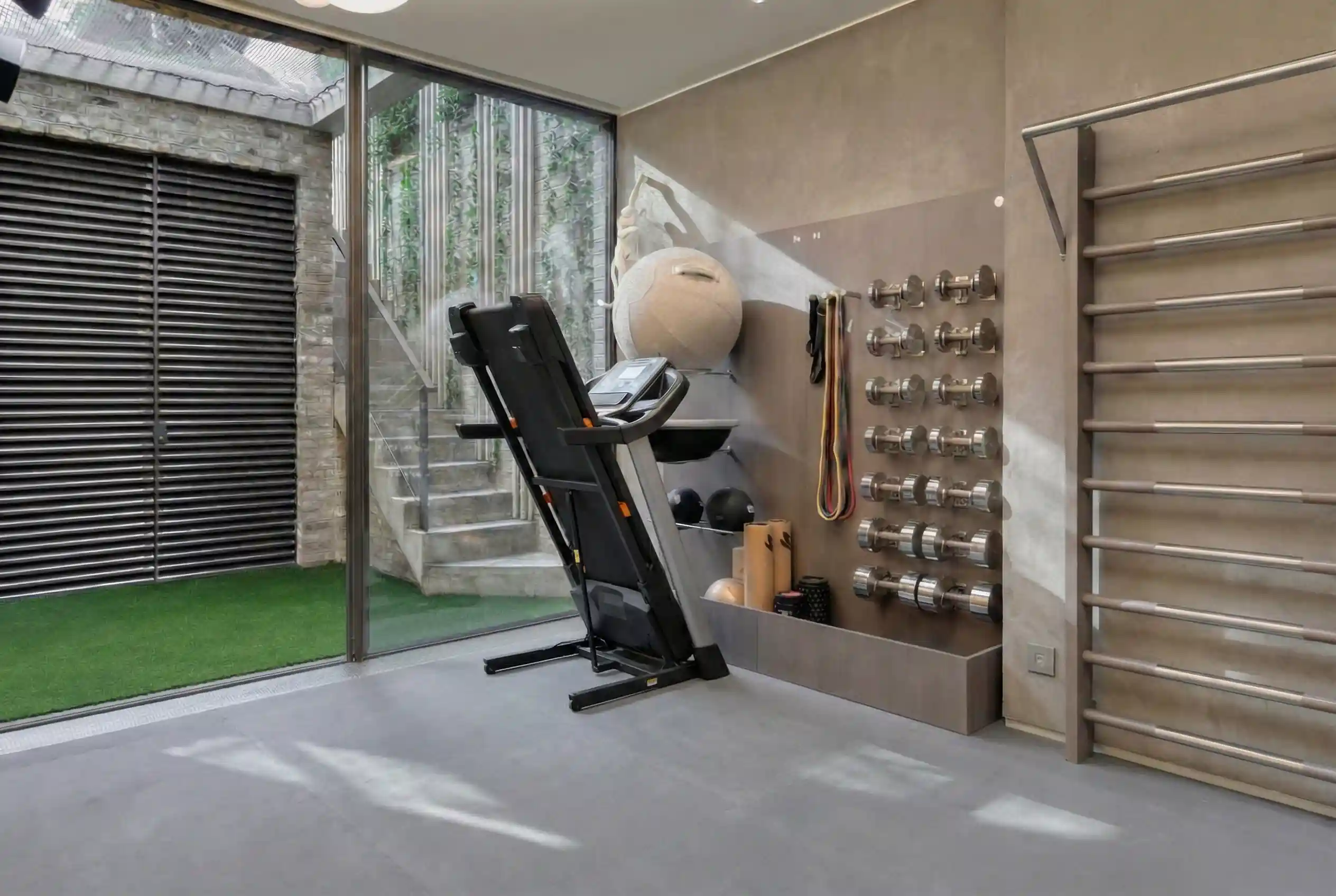 NordicTrack T Series 6.5s treadmill in folded upright position in modern home gym with artificial turf flooring, wall-mounted chrome dumbbell rack, stall bars, and glass door to outdoor courtyard