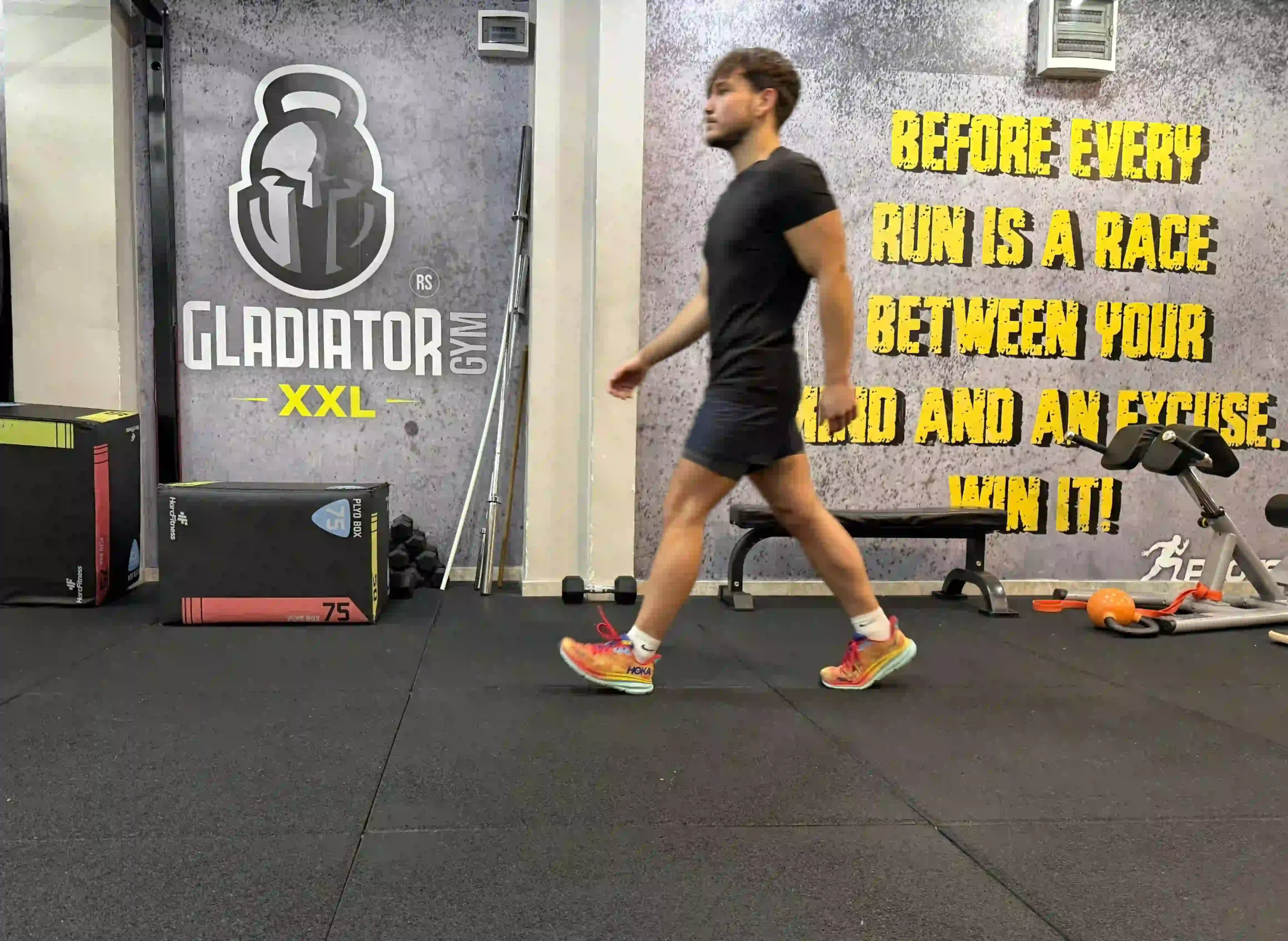 A man in athletic wear walking across a gym floor, demonstrating a natural walking gait with a heel strike.