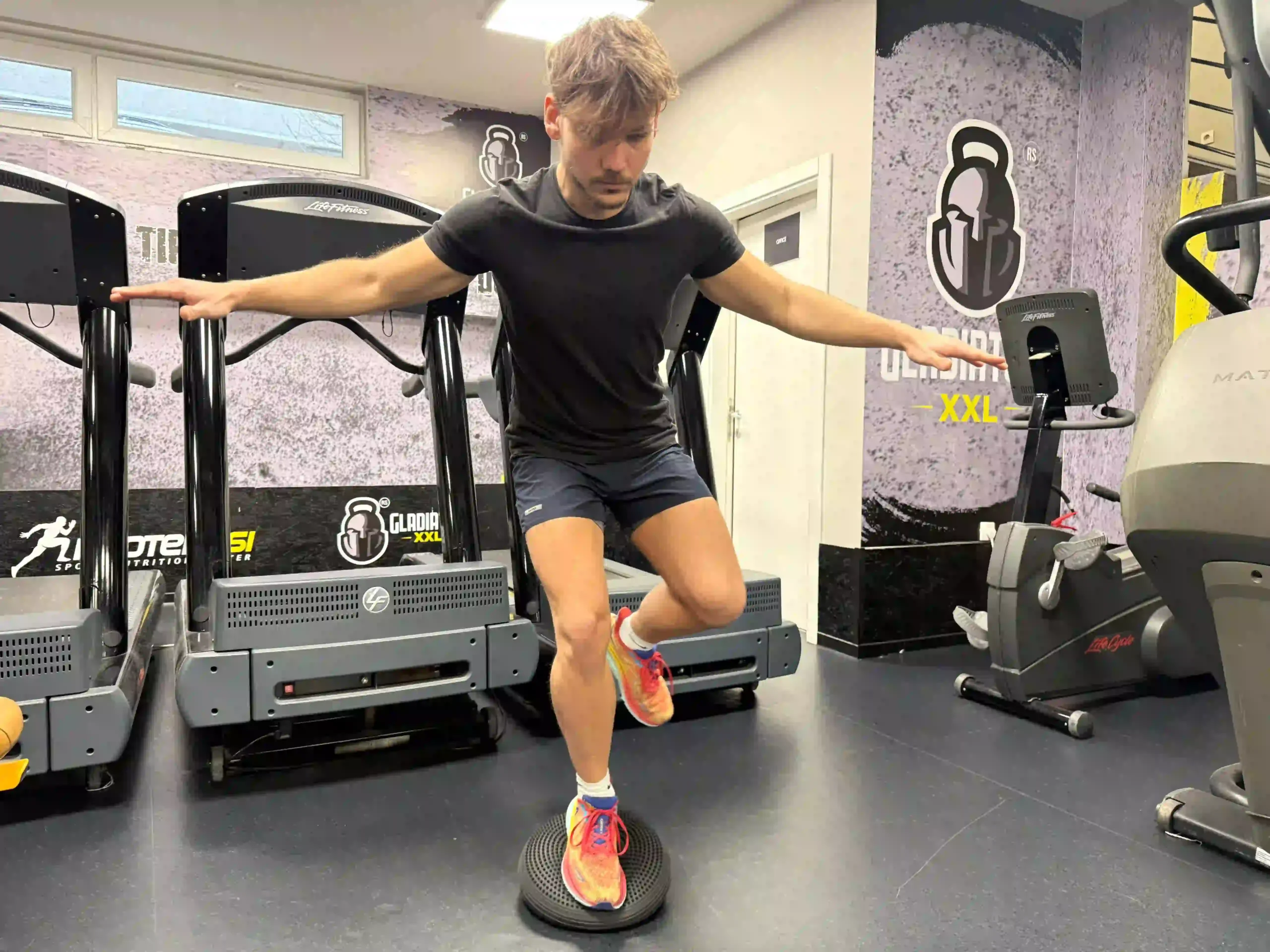 A young man in athletic wear performing a one-leg balance exercise on a black BOSU ball in a gym.
