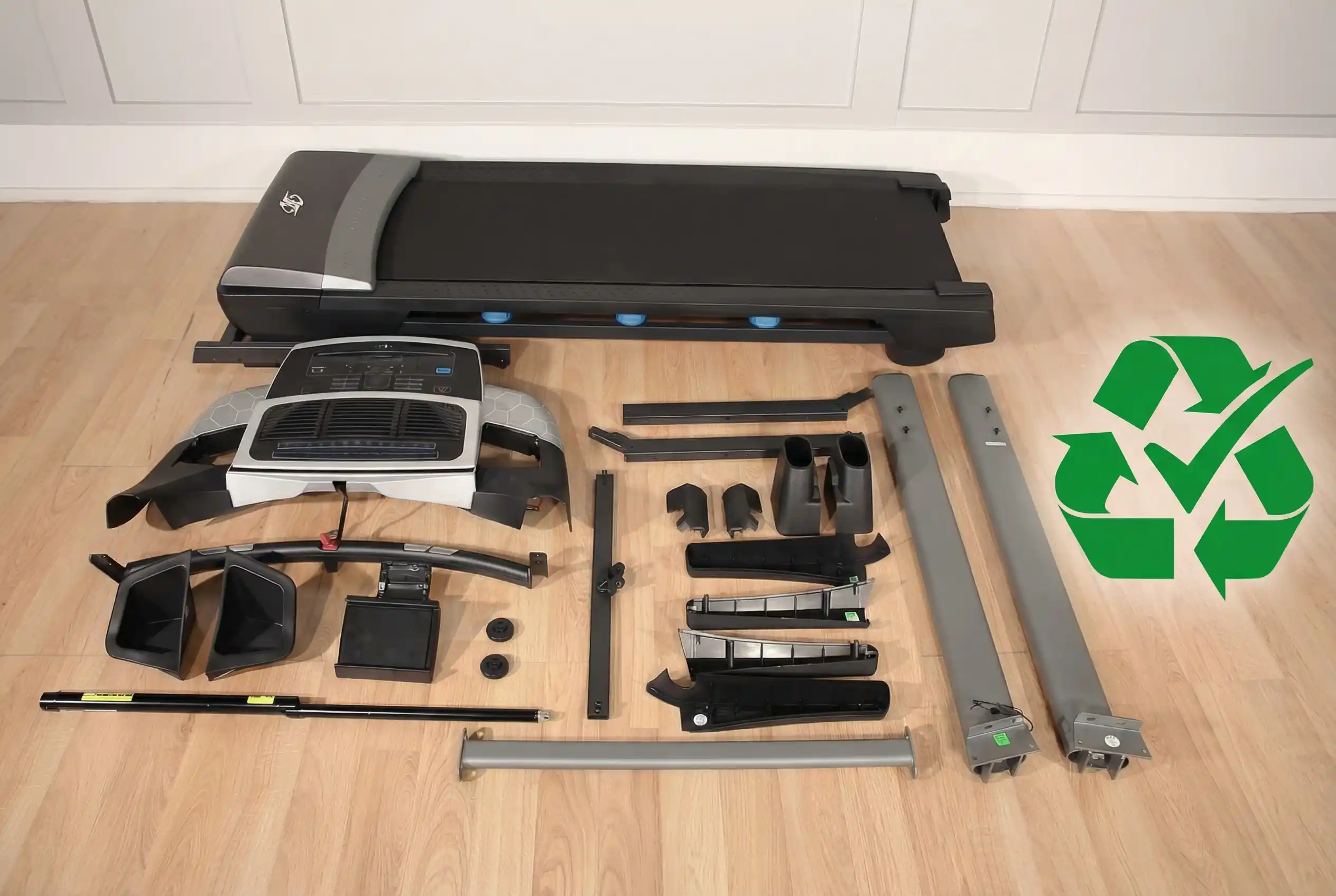 Completely disassembled treadmill with all component parts organized on hardwood floor including main running deck, console, upright posts, motor covers, frame pieces, and hardware, with green recycling symbol and checkmark overlay indicating recyclable materials