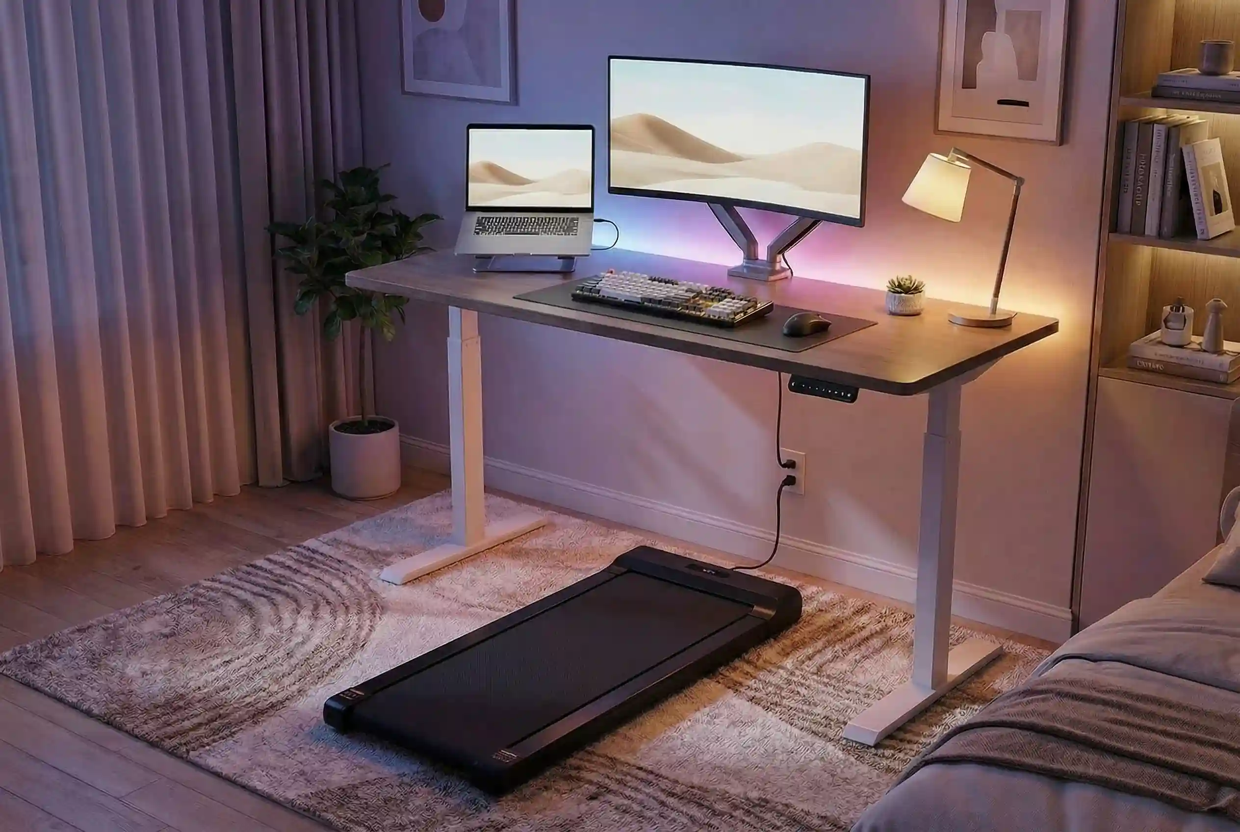 Sperax Walking Pad positioned under electric standing desk with dual monitor setup, RGB desk lighting, indoor plants, and modern home office decor in evening ambient lighting