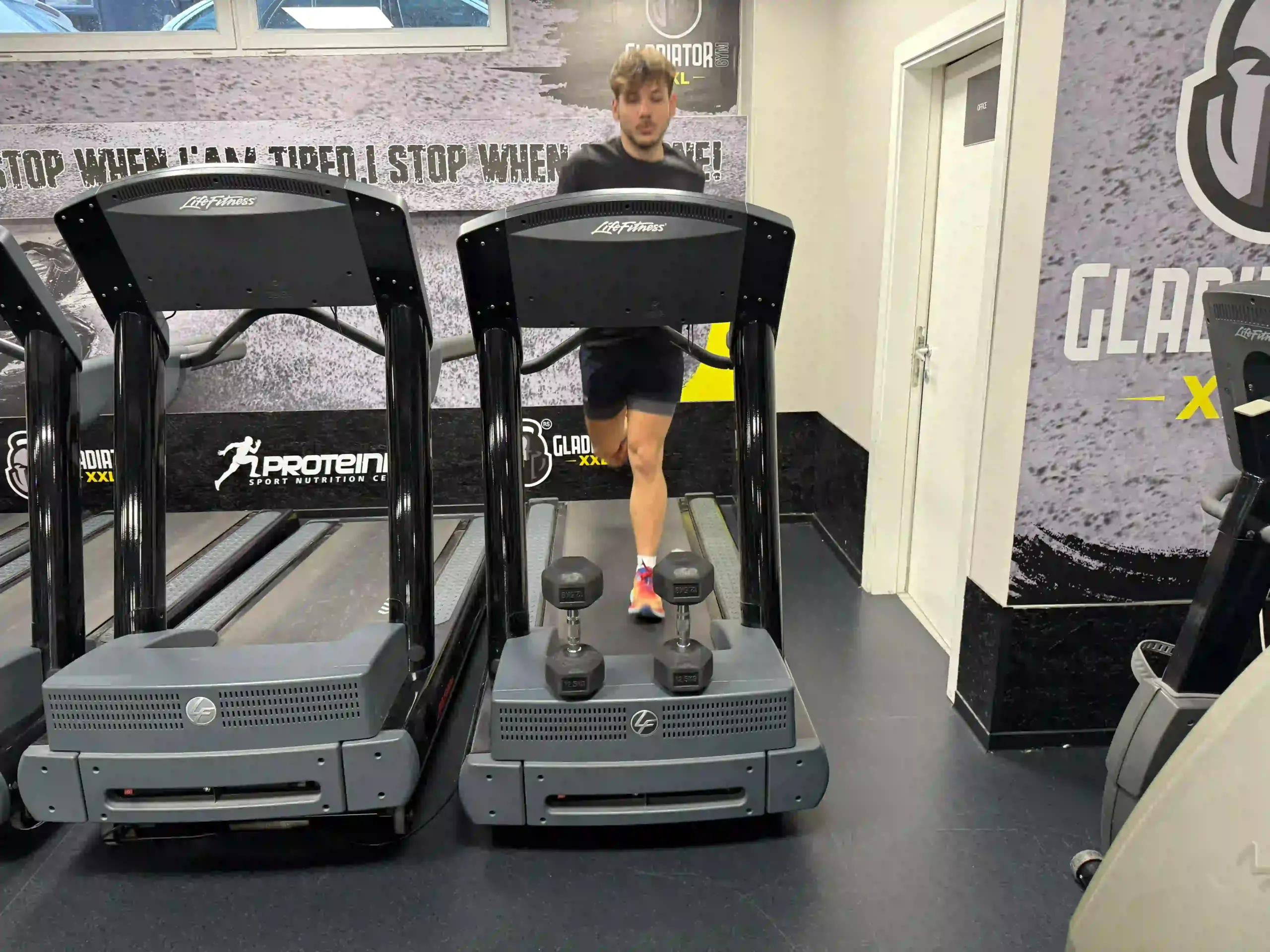 Milutin walking on commercial treadmill with dumbbells placed on treadmill base in dark shorts and colorful running shoes at gym facility