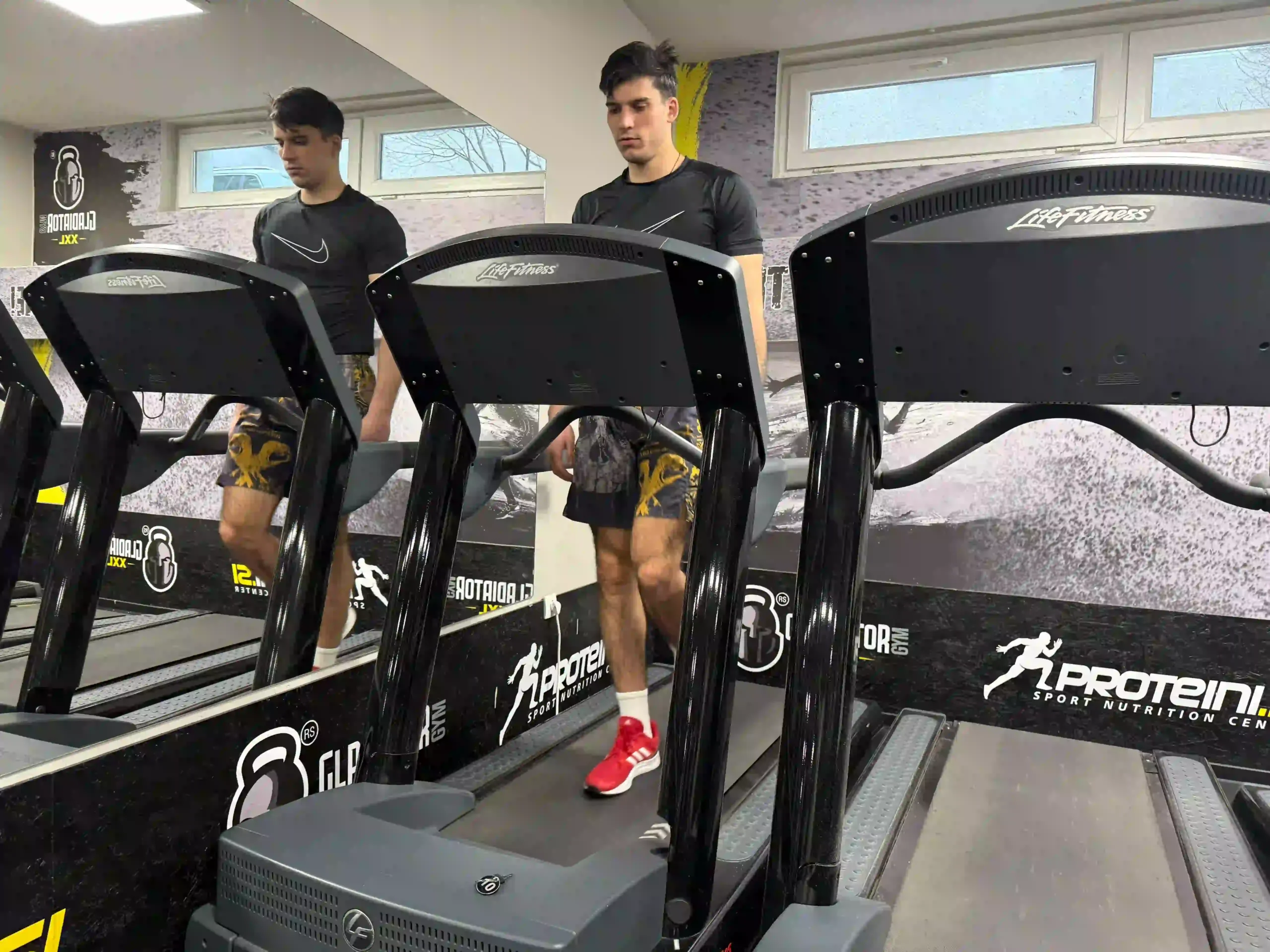 A young man named Vanja wearing a black t-shirt and red sneakers, walking on a treadmill in front of a mirror at the gym.