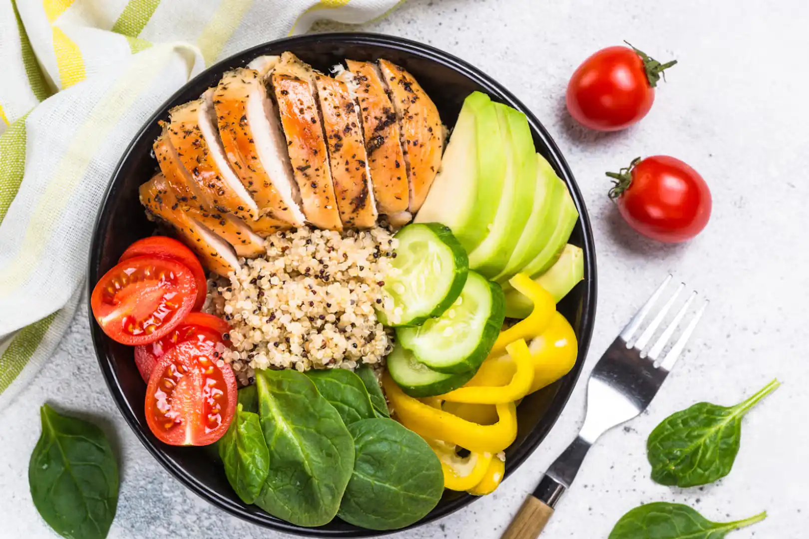 A healthy fitness meal bowl featuring grilled chicken breast, quinoa, sliced avocado, spinach, cucumbers, and cherry tomatoes.