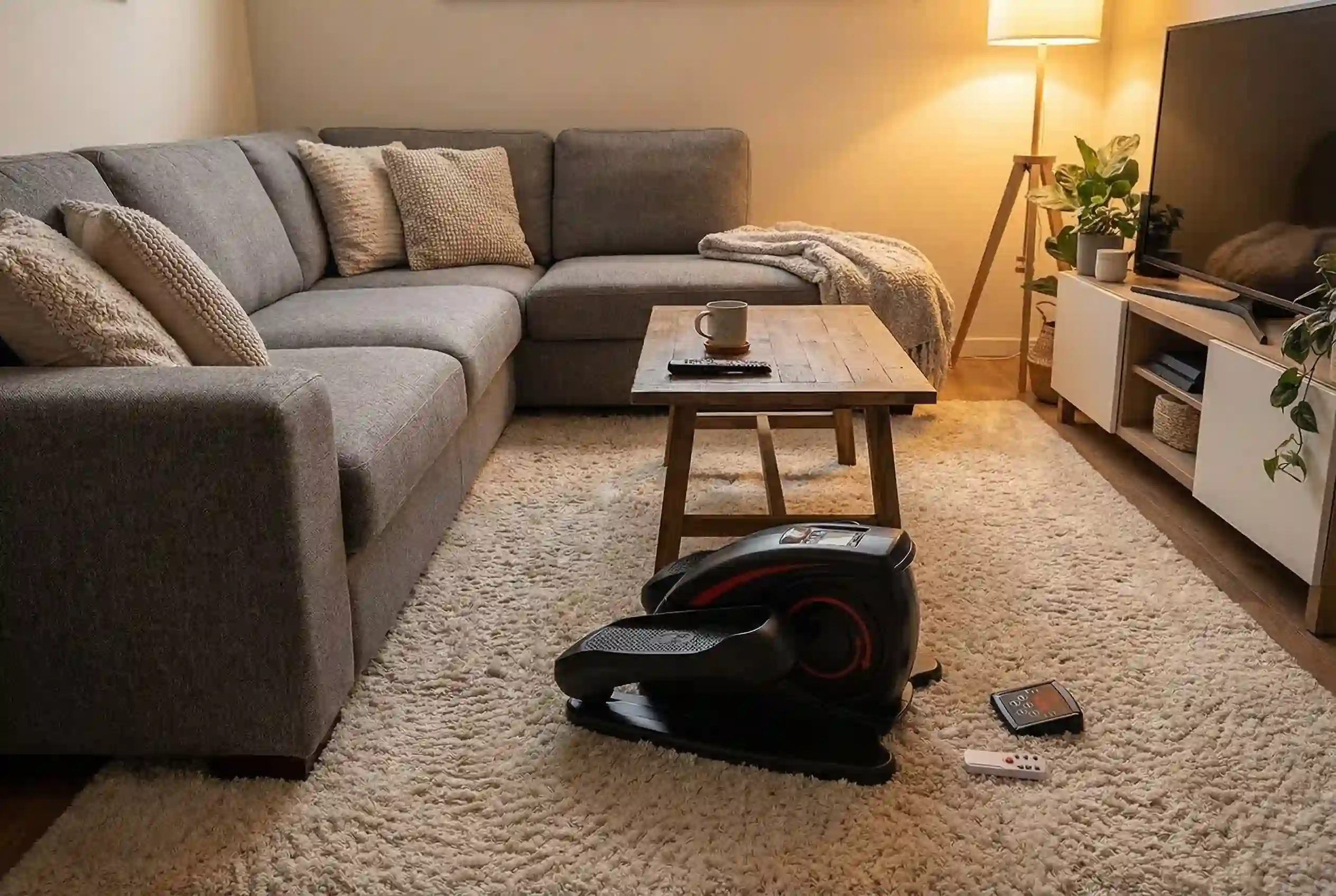 Ancheer under-desk elliptical with red accent ring and remote control on cream shag rug in cozy living room with gray sectional sofa, knit throw blanket, rustic wood coffee table, floor lamp, indoor plants, and wall-mounted TV
