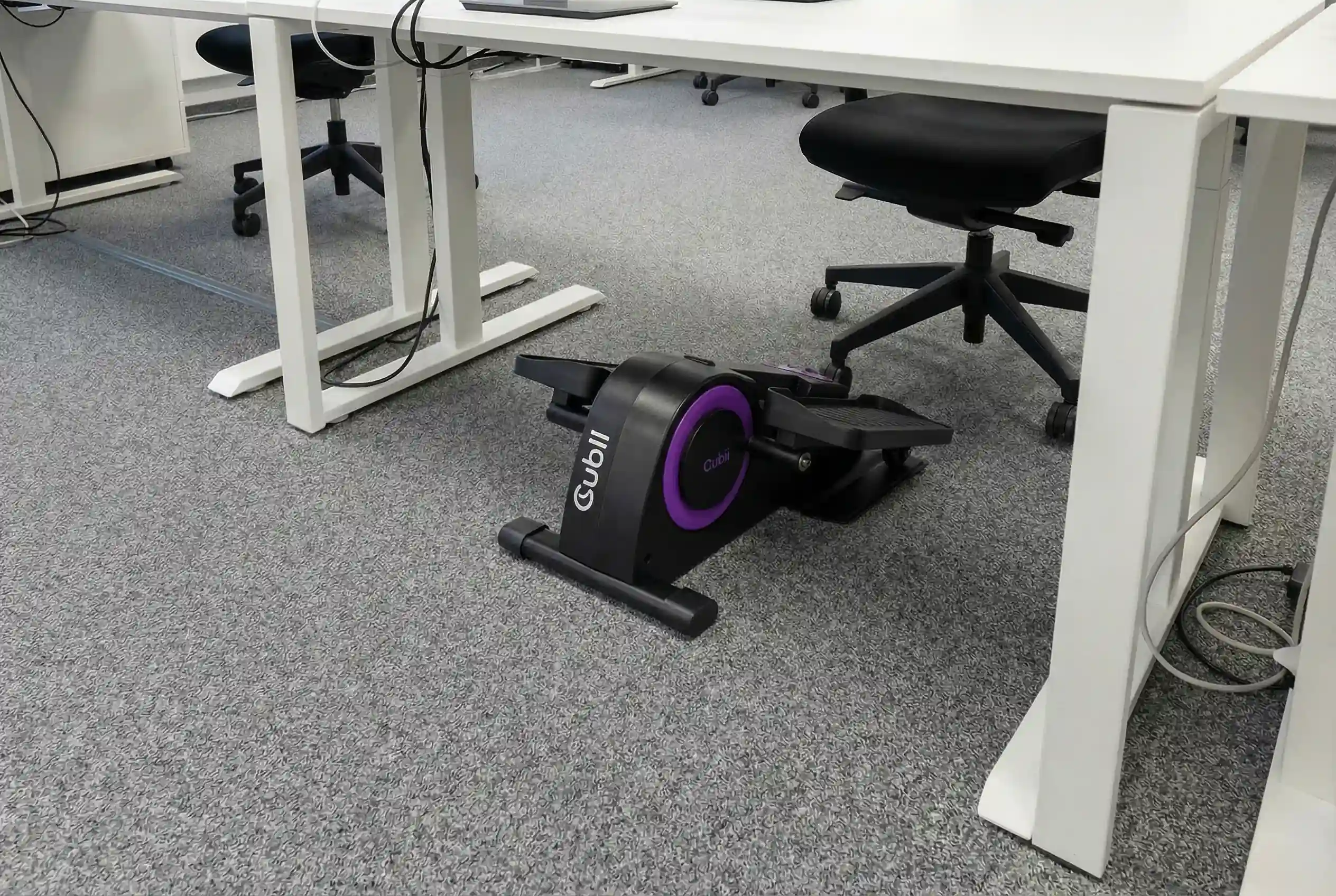 Cubii-JR1 Cubii JR1 compact seated elliptical pedal exerciser with purple accent ring positioned on gray carpet under white office desk with black office chair in modern workspace