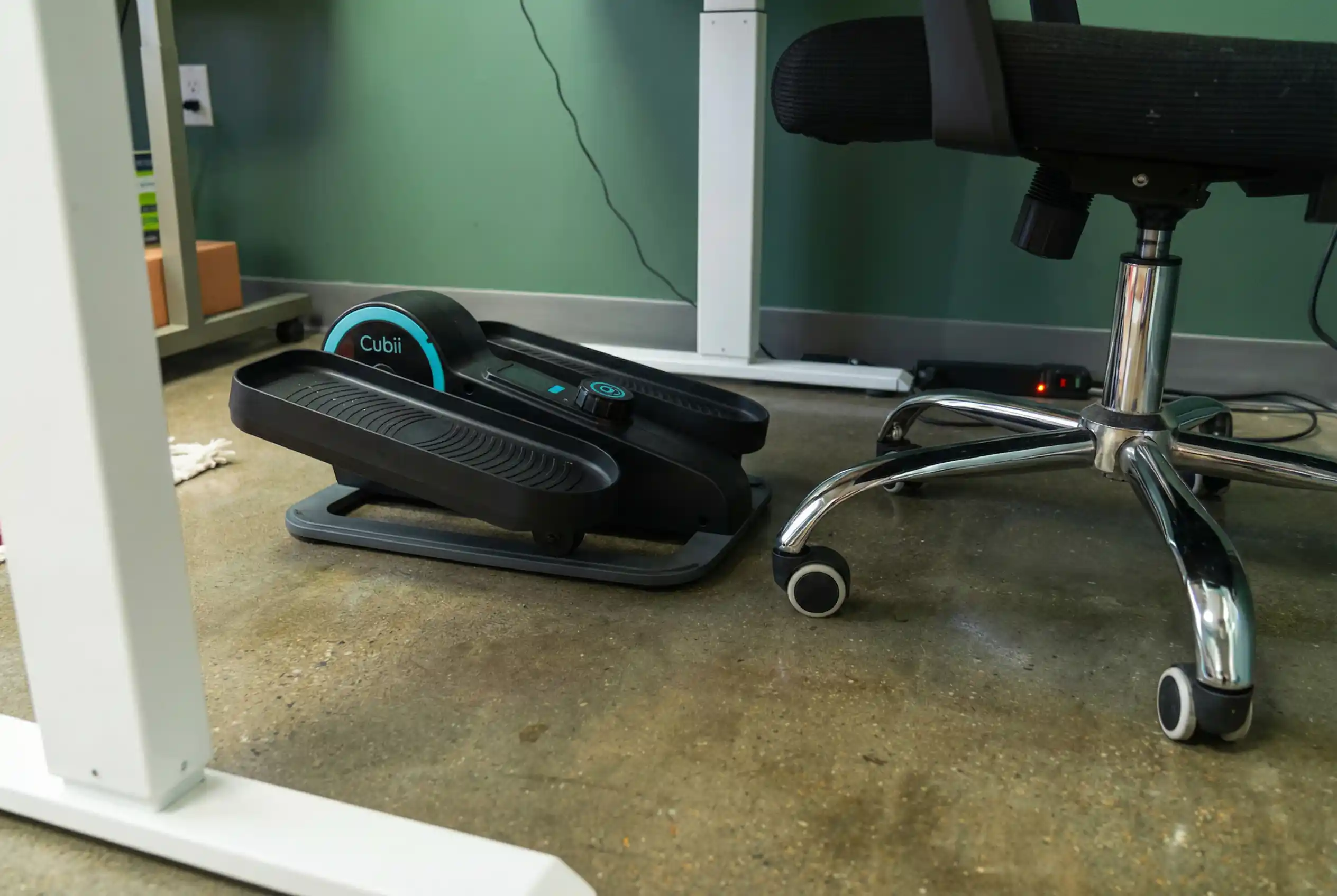 Cubii-Move Cubii Move compact under-desk elliptical pedal exerciser with blue accent ring and digital display positioned under office desk on polished concrete floor with chrome chair and green walls