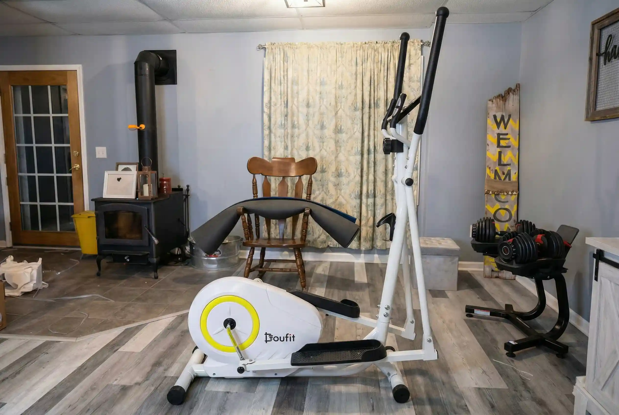 Image (Doufit-EM-01.webp):
"Doufit EM-01 elliptical machine with white frame and yellow flywheel accent on vinyl plank flooring in basement home gym with black wood-burning stove, wooden rocking chair, floral curtains, adjustable dumbbell set on stand, and rustic welcome sign on gray walls