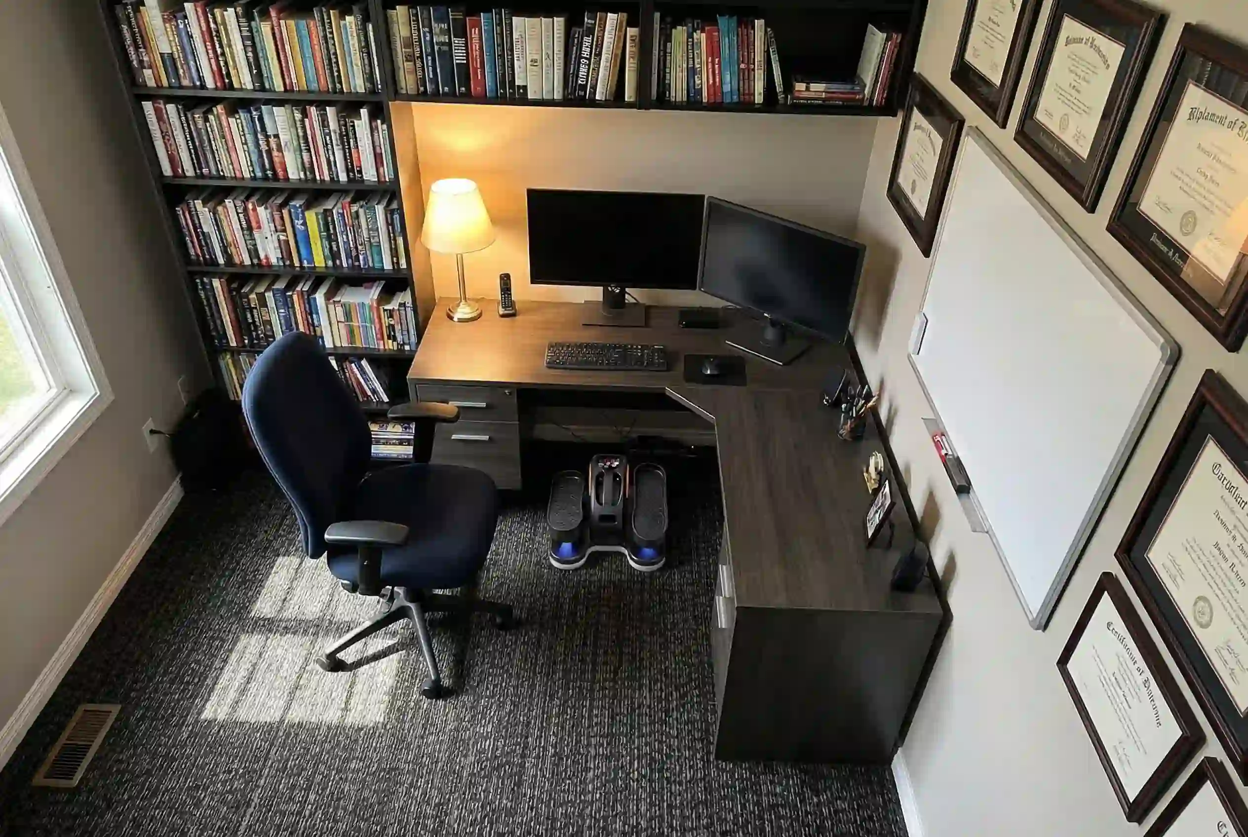 GUGTTR under-desk elliptical with blue accent lights positioned beneath L-shaped dark walnut desk with dual monitor setup, keyboard, desk lamp, navy ergonomic office chair, floor-to-ceiling bookshelves, and framed certificates on wall