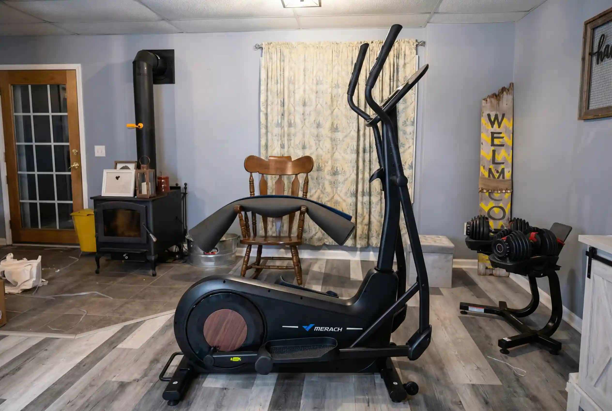 Merach-E27 MERACH E27 elliptical machine with moving handlebars and copper-colored flywheel in basement home gym with blue walls, wood stove, wooden chair, patterned curtains, adjustable dumbbells, and laminate flooring