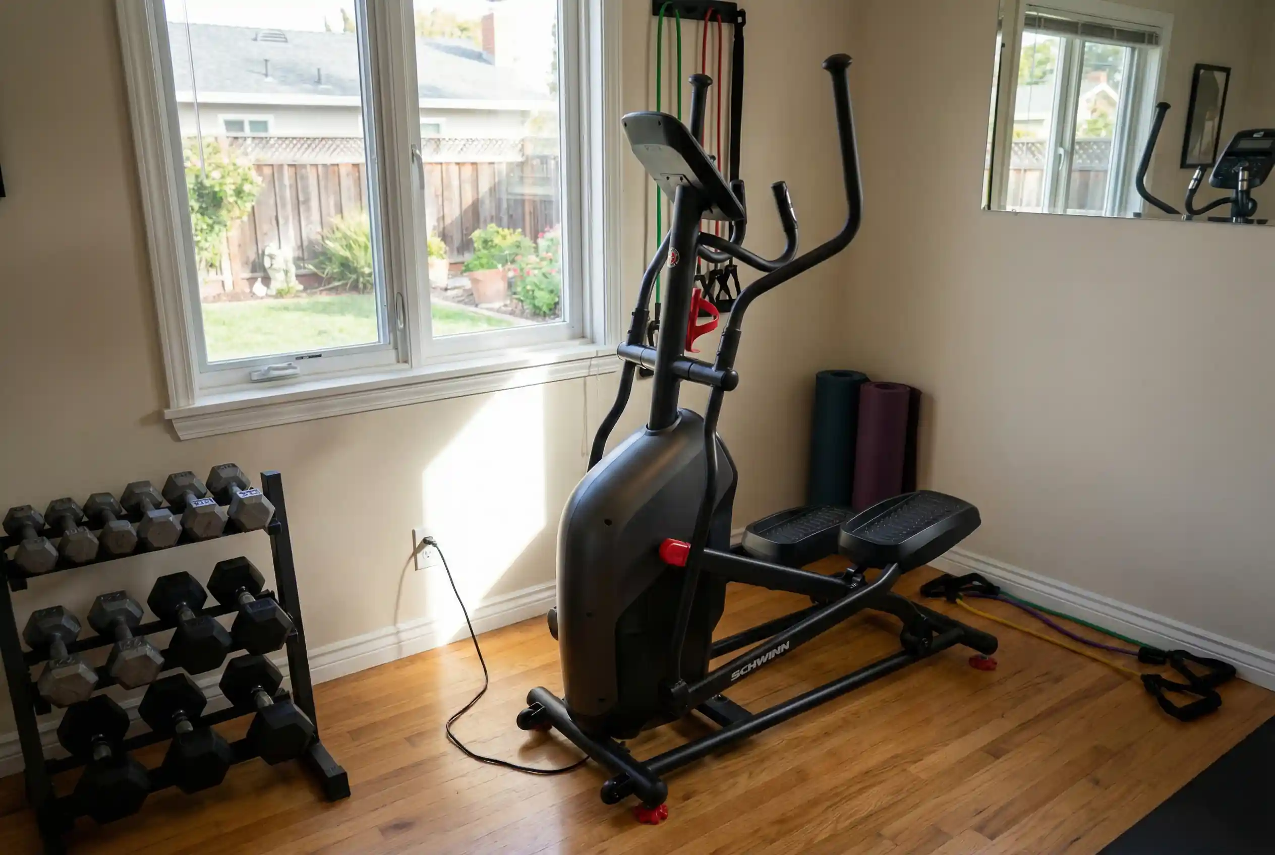 Schwinn-411 Schwinn 411 compact elliptical machine with moving handlebars and red accent features on hardwood floor in bright home gym with window overlooking backyard, dumbbell rack, resistance bands on wall, and foam rollers