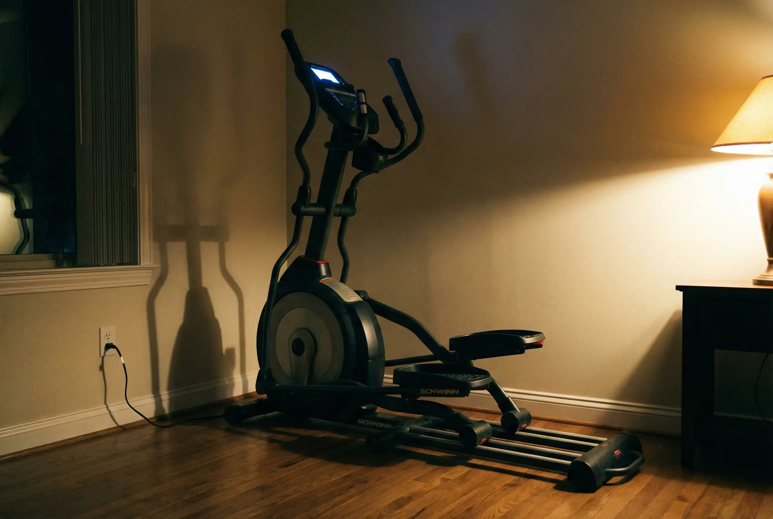 Schwinn-470 Schwinn 470 elliptical machine with illuminated digital console and moving handlebars on hardwood floor in evening home setting with window, warm lamp lighting, and beige walls