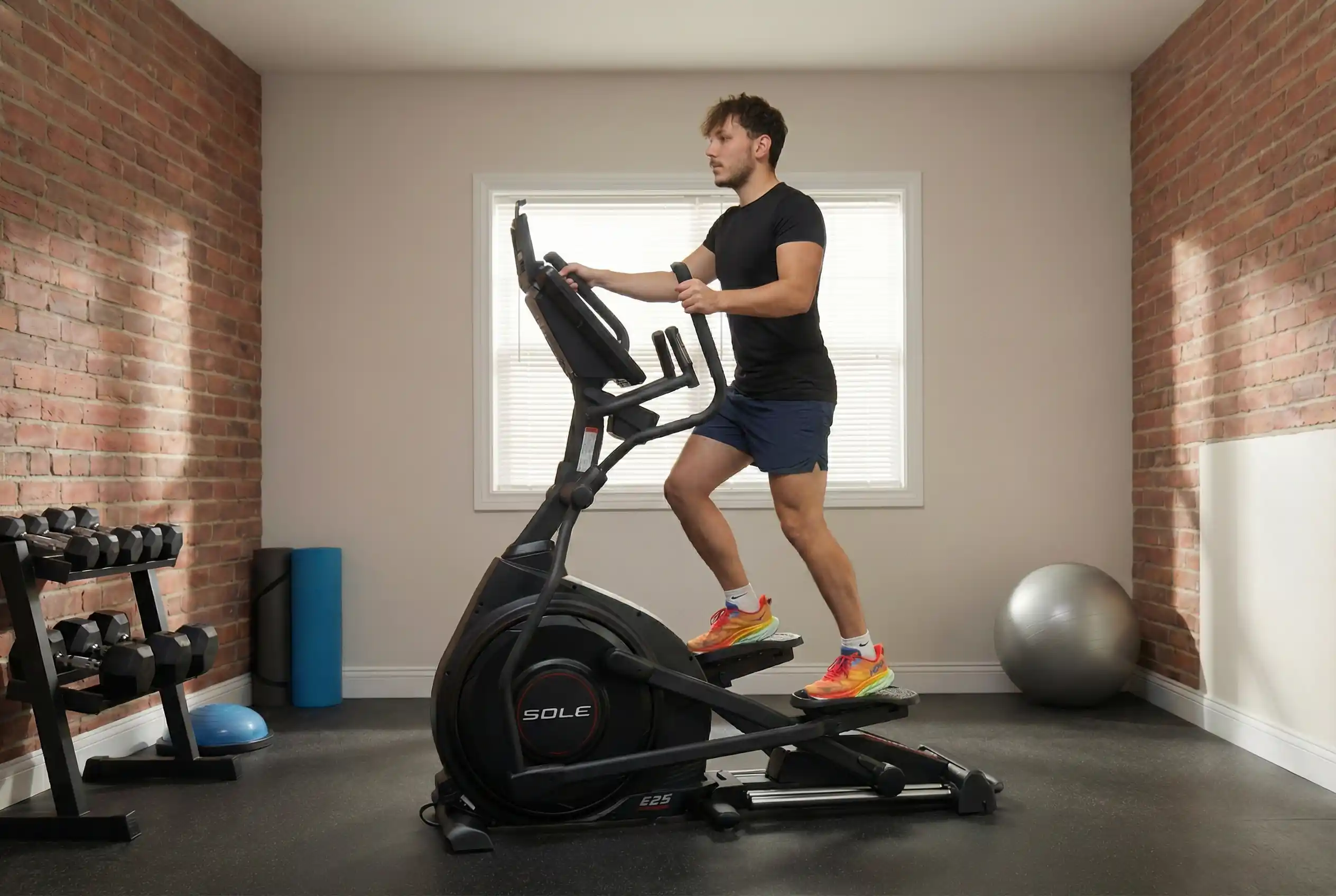 Sole-E25-Testing Milutin exercising on Sole E25 elliptical machine in black athletic shirt, blue shorts, and orange running shoes in home gym with exposed brick walls, natural window lighting, dumbbell rack, stability ball, and foam roller