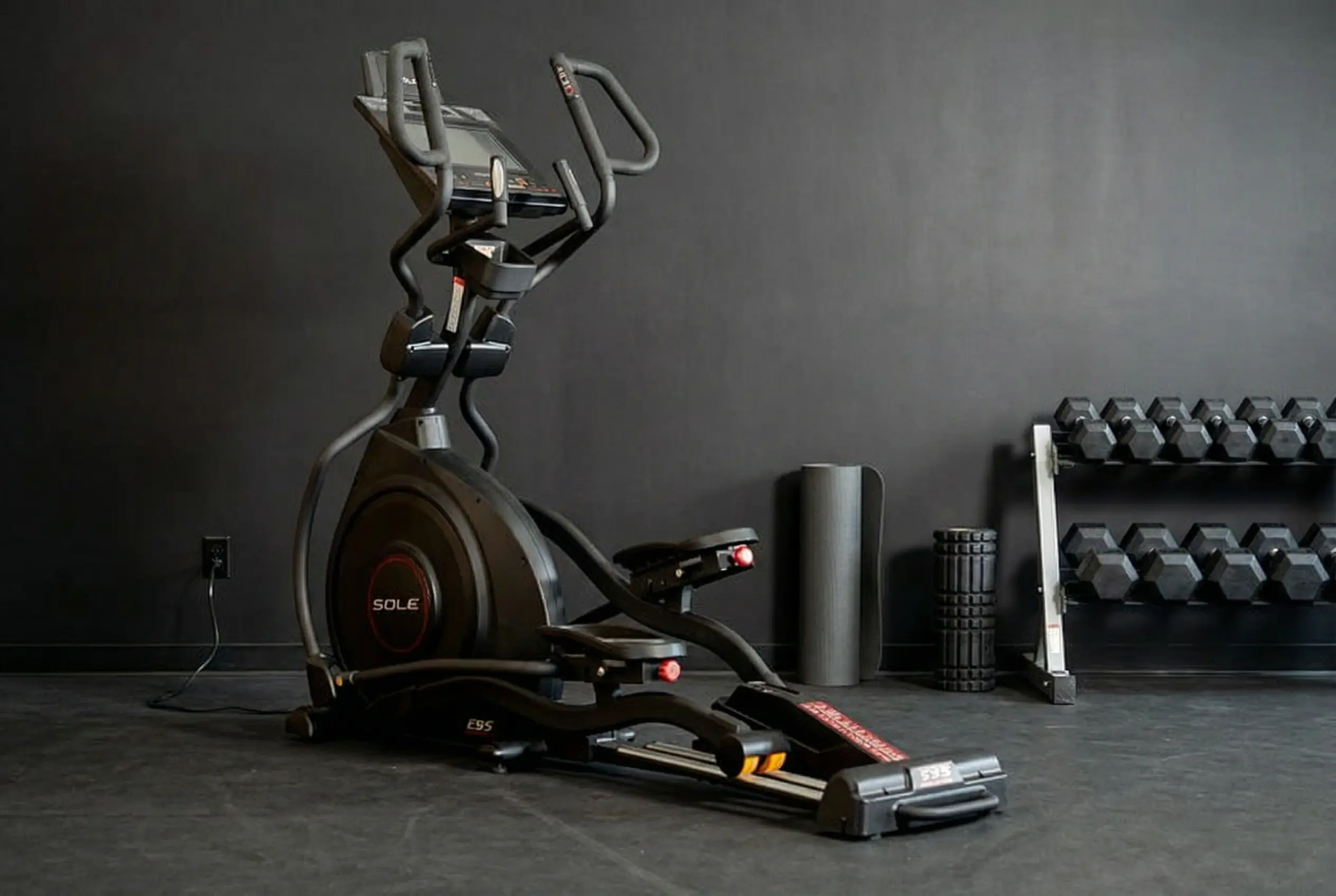 Sole-E95 Sole E95 elliptical machine with digital console, moving handlebars, and large flywheel in gym setting with dark gray walls, dumbbell rack, and foam rollers