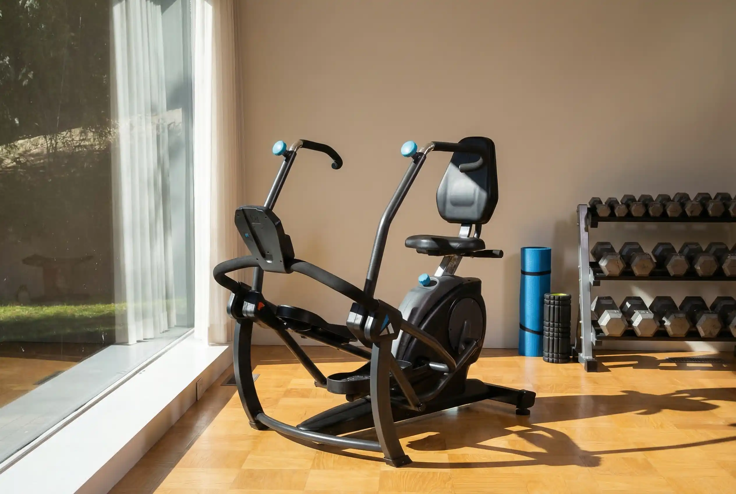 Teeter-Freestep-LT1 Teeter Freestep LT1 recumbent cross trainer with padded seat, blue accent grips, and curved frame design on hardwood floor in home gym with floor-to-ceiling window, natural lighting, dumbbell rack, foam roller, and beige walls