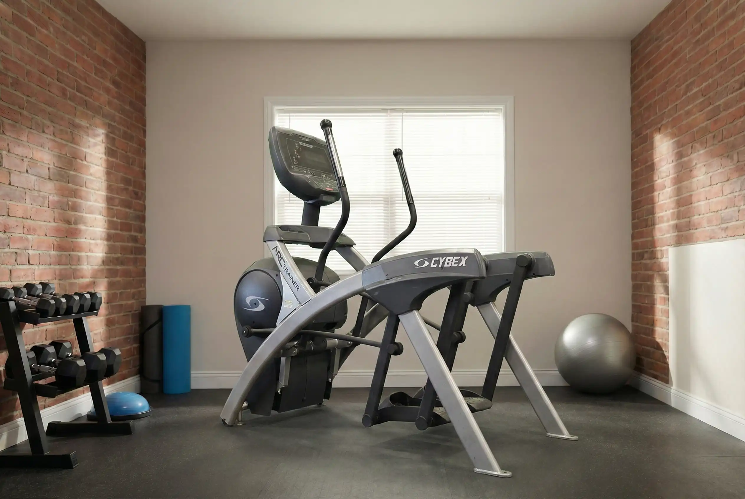 Cybex Arc Trainer placed in a home gym environment, with a visible gym ball, dumbell rack.