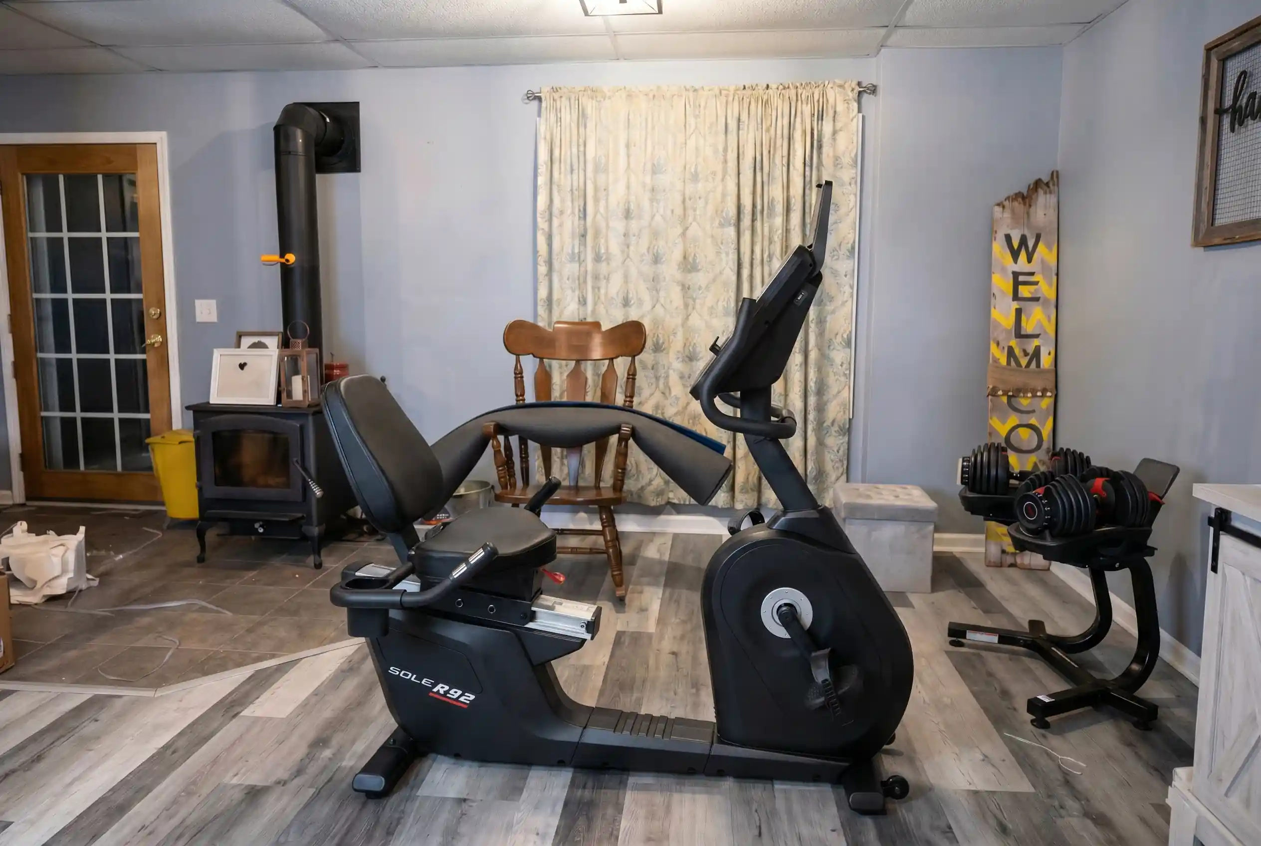 Sole R92 recumbent exercise bike with large touchscreen display and cushioned seat, placed in a home gym space alongside adjustable dumbbells on a stand