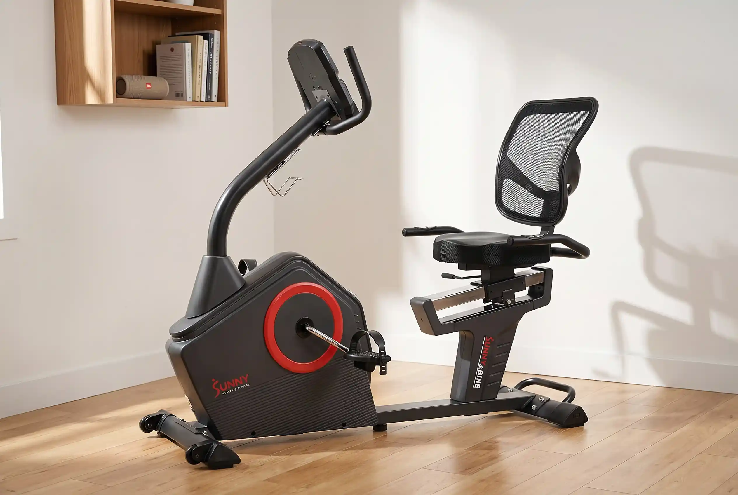 Sunny Health & Fitness recumbent exercise bike with mesh backrest and red flywheel accent, placed on hardwood floor next to a wooden bookshelf in a bright minimalist room