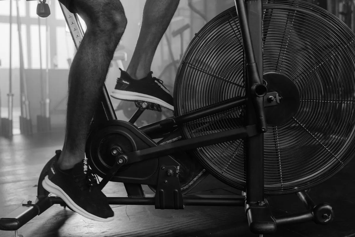 Black and white close-up of a person's legs on an air bike with a large fan wheel during a gym workout