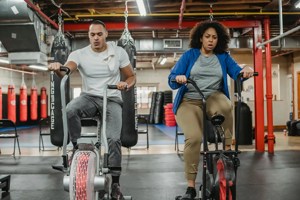 Man and woman pushing hard on air bikes in a gym with heavy bags in the background during a high intensity session