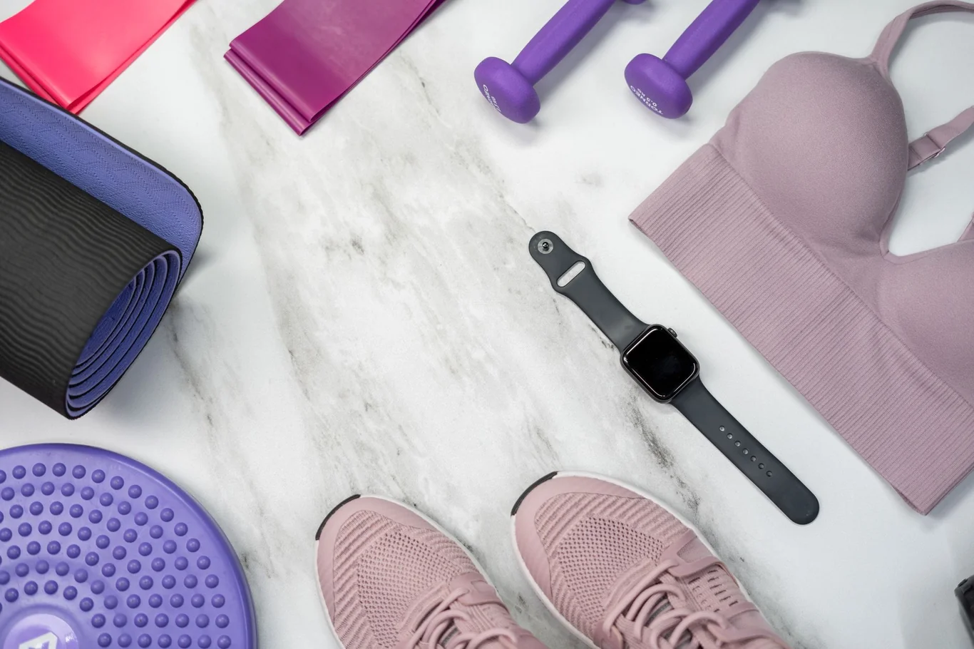 Black smartwatch on a marble surface surrounded by dumbbells, resistance bands, sneakers, and a sports bra