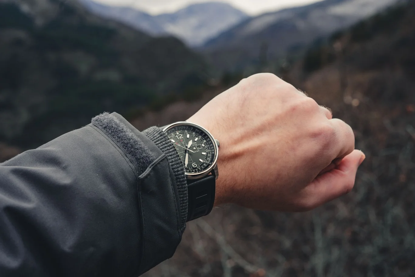 Black sport watch on a wrist with a hiking jacket sleeve and mountain backdrop
