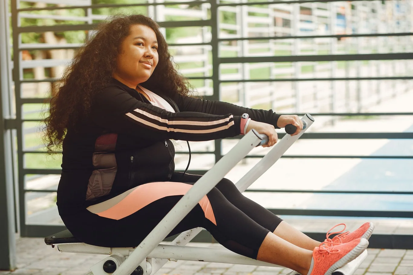 Woman with knees bent and arms extended, gripping the handle on a rowing machine outdoors