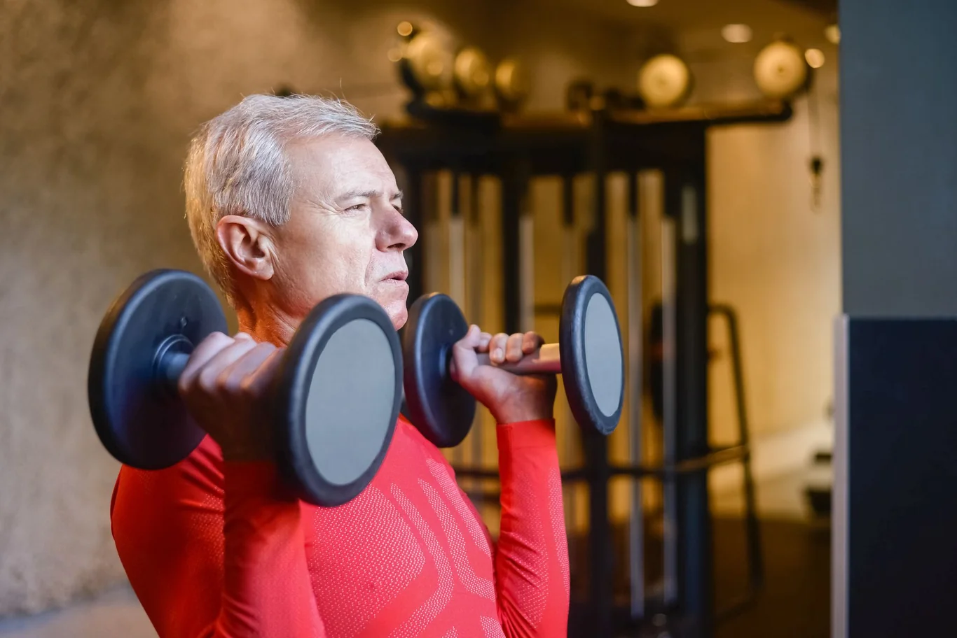 Older man in a red long sleeve shirt doing dumbbell curls at the gym