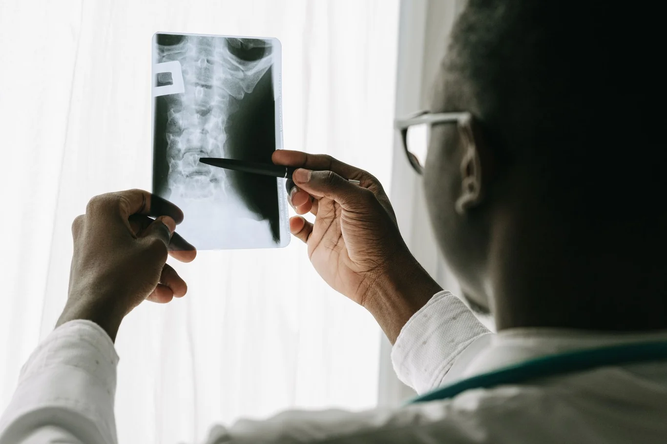 Doctor holding a spinal X-ray up to the light while pointing at a vertebra with a pen