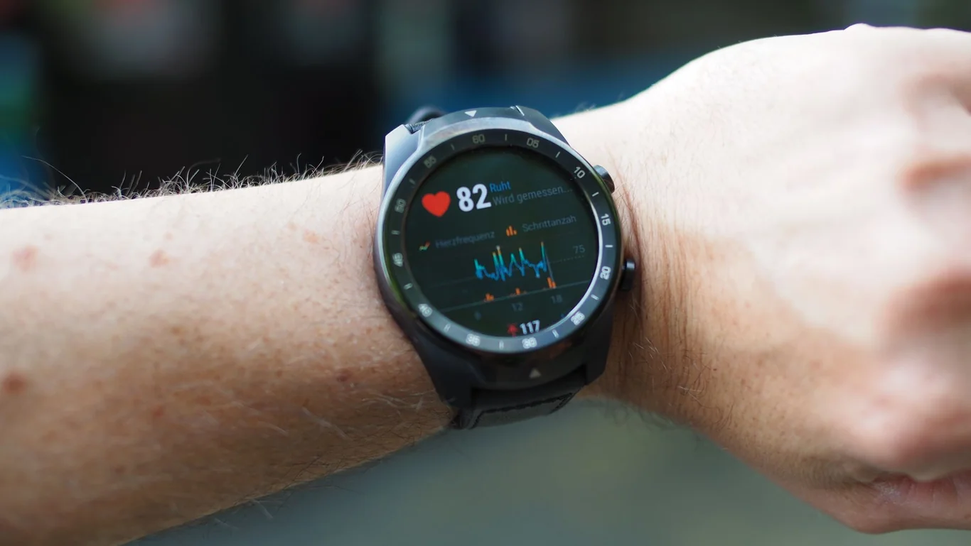 Round smartwatch on a wrist showing a heart rate reading of 82 with a line graph below