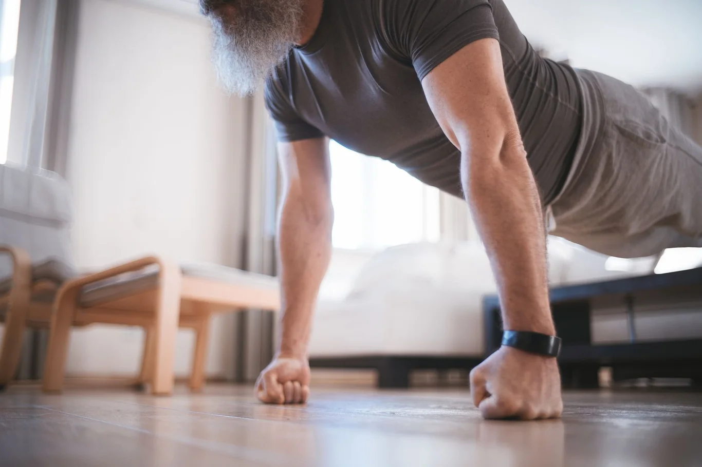 Older bearded man doing a push-up on a wood floor while wearing a black fitness tracker