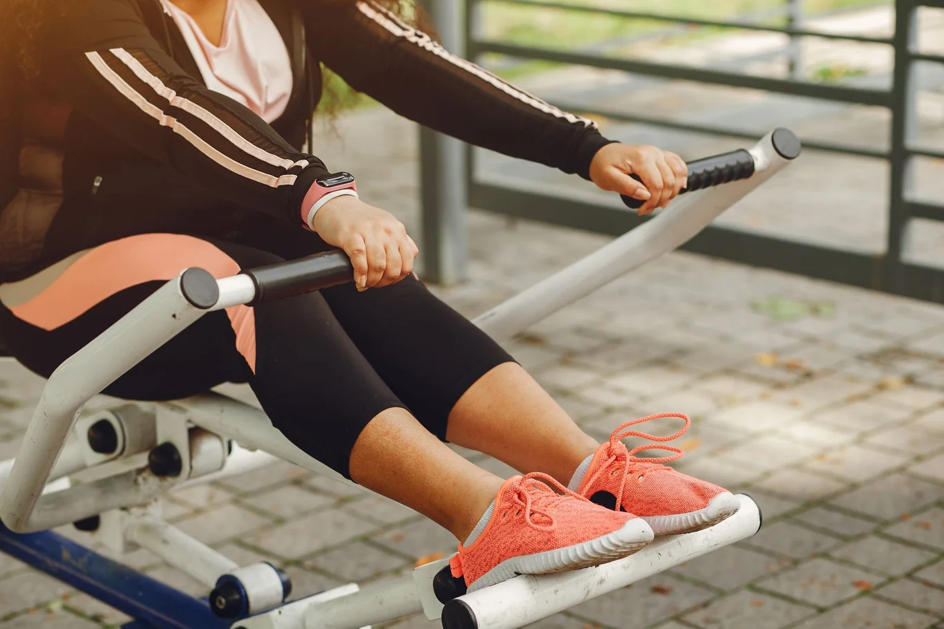 Woman in a black tracksuit using a rowing machine outdoors with orange sneakers
