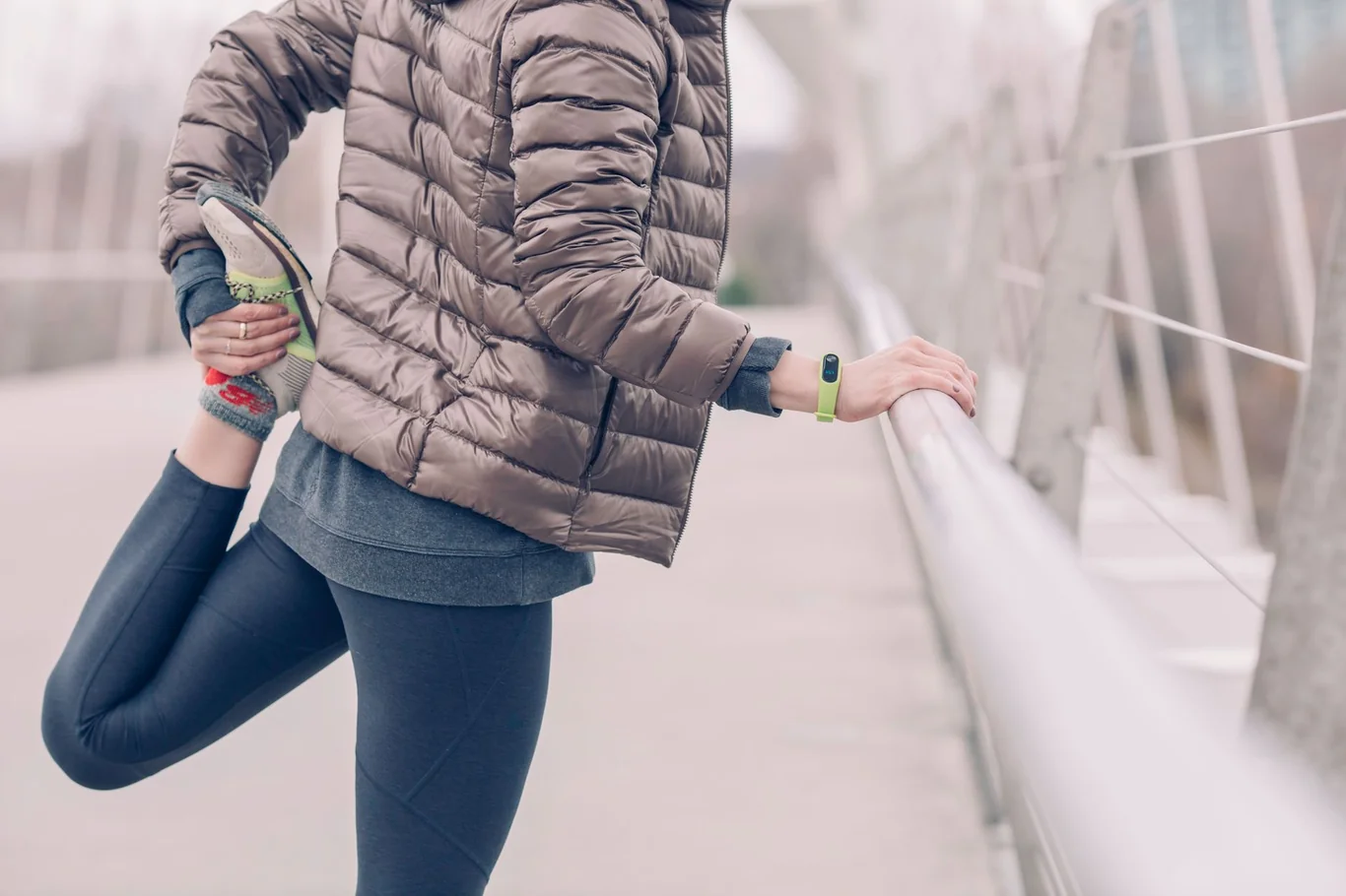 Woman in a puffer jacket stretching her leg outdoors while wearing a green fitness tracker