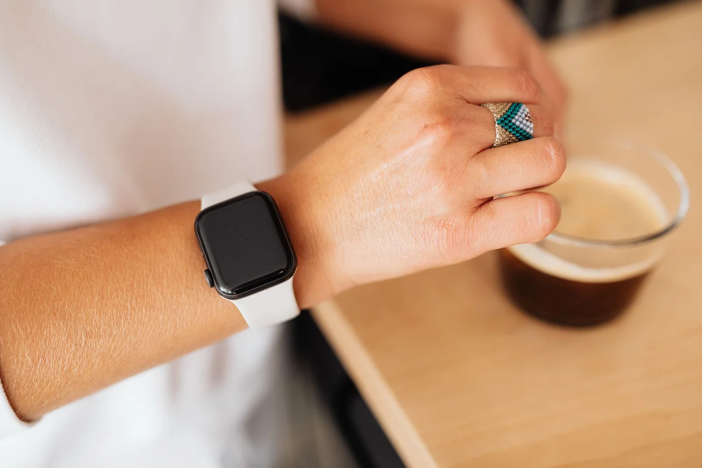 Person wearing a white band smartwatch on their wrist beside a glass of iced coffee on a wooden table