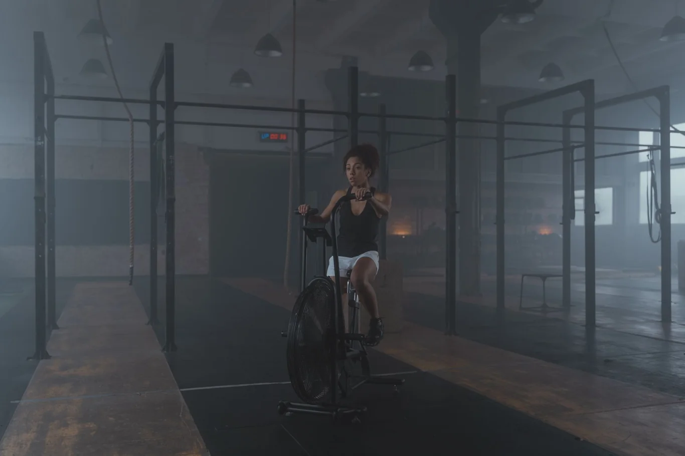 Woman pedaling an air resistance bike in a dim industrial-style gym with a pull-up rig