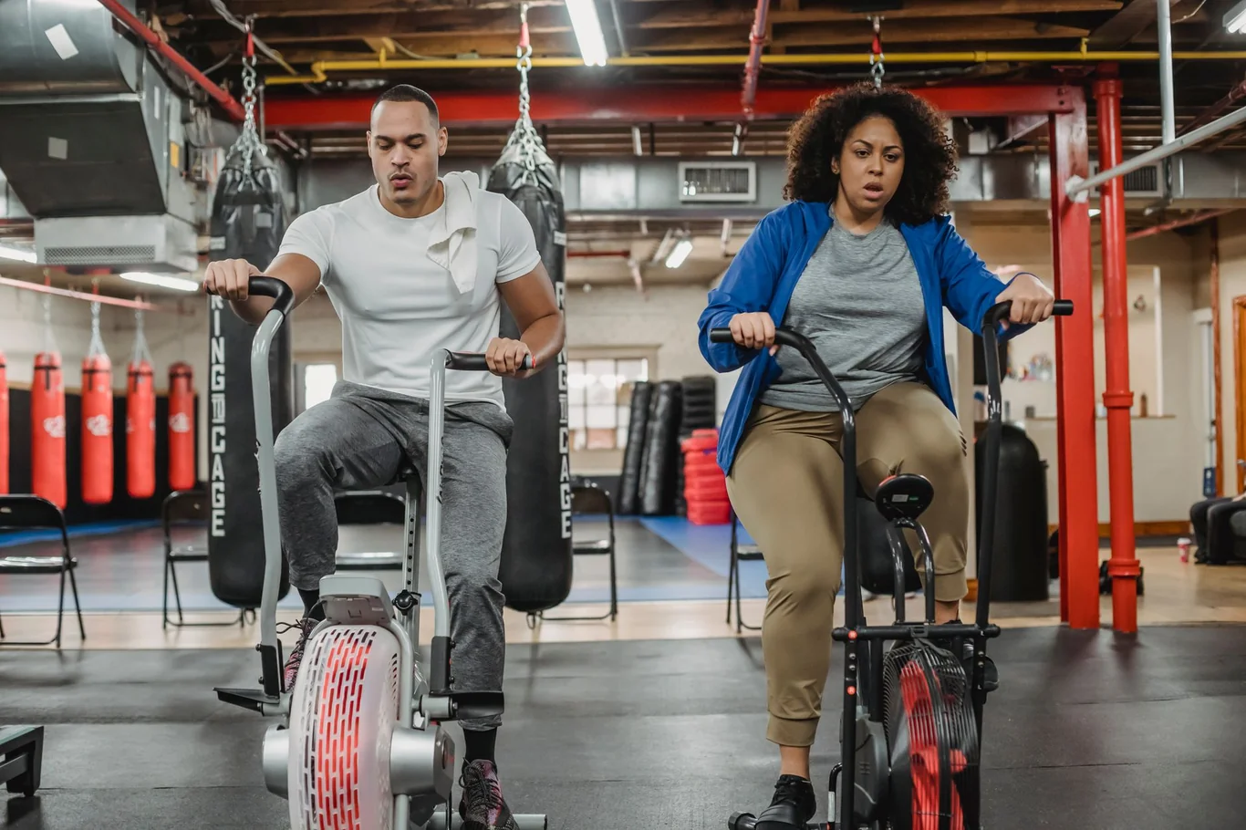 Man and woman pedaling air bikes together in a bright gym with boxing bags