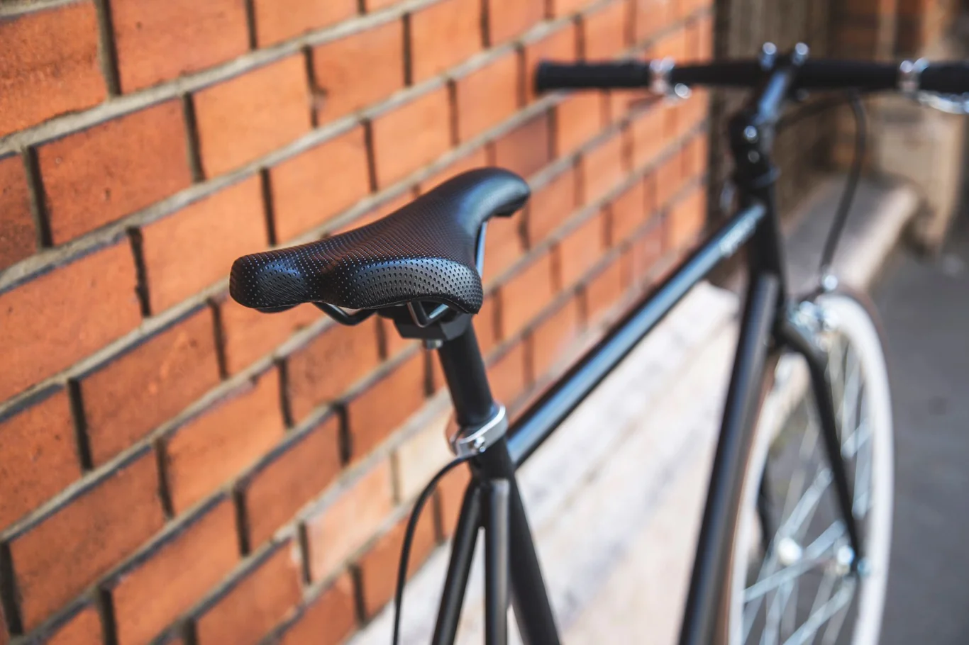 Black bicycle saddle mounted on a frame near a brick wall with drop handlebars visible