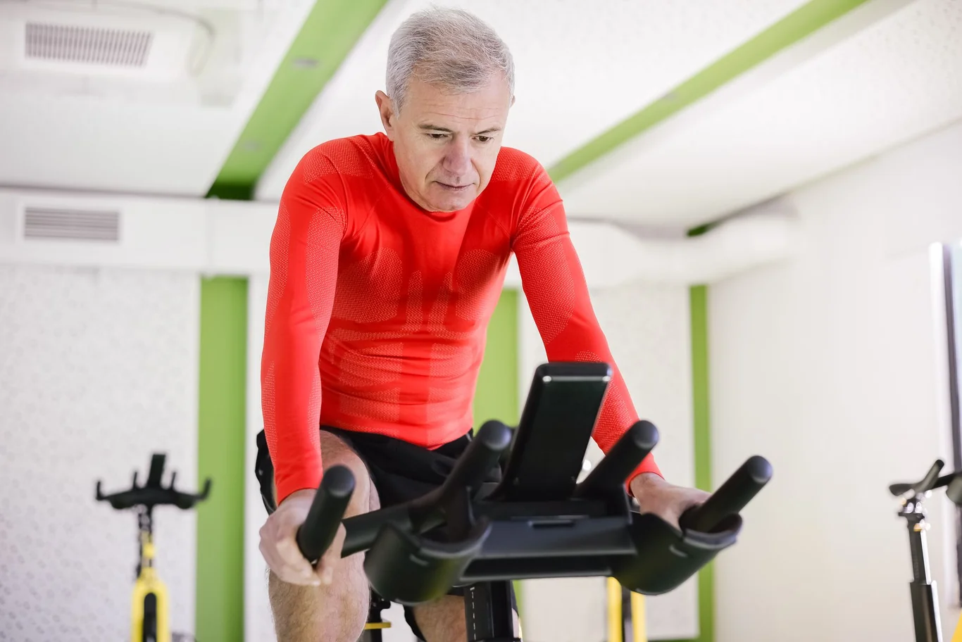 Older man in a red shirt cycling on an indoor stationary bike while checking his phone