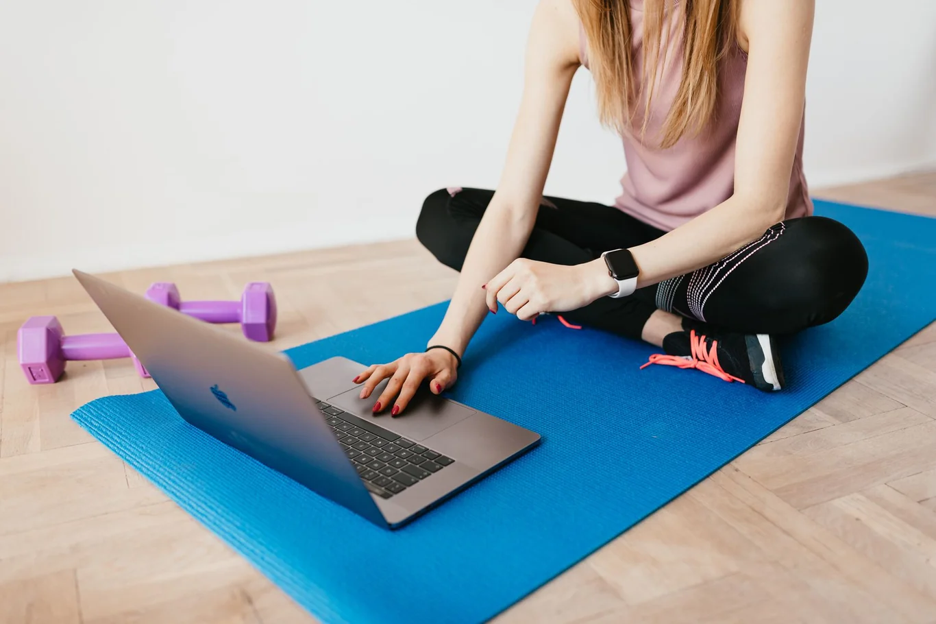 Woman on a yoga mat wearing a smartwatch beside a laptop and dumbbells