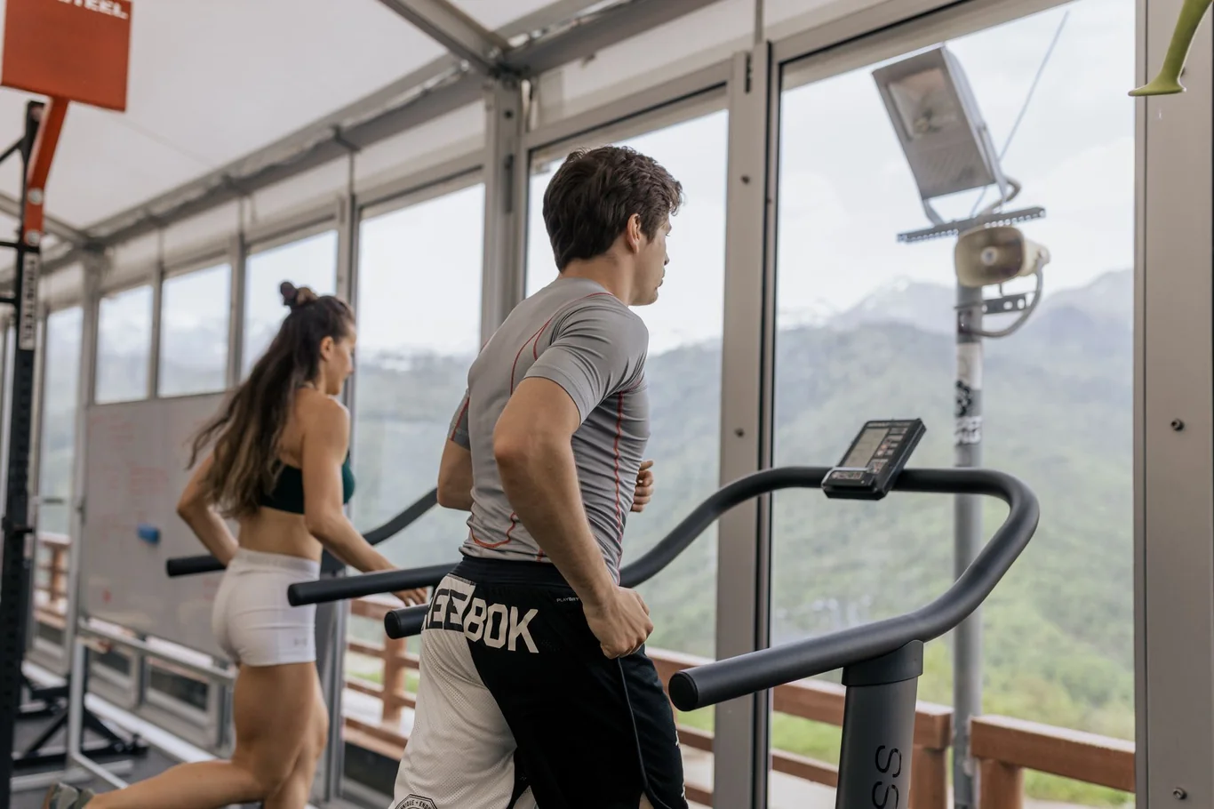 Man running on a treadmill next to a woman working out in a gym with large windows